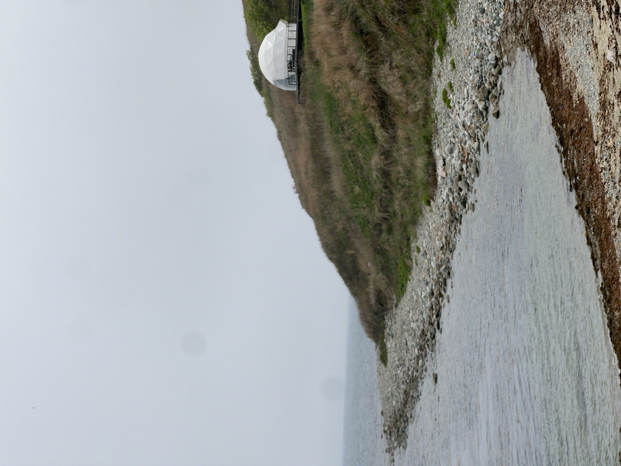 A tilted long shot reveals a scene featuring a body of water, a pebble beach, and a grassy cliff. A white geodesic dome-like structure is perched on the cliff. The sky is overcast. The scene appears to be in Zarubino, Russia.