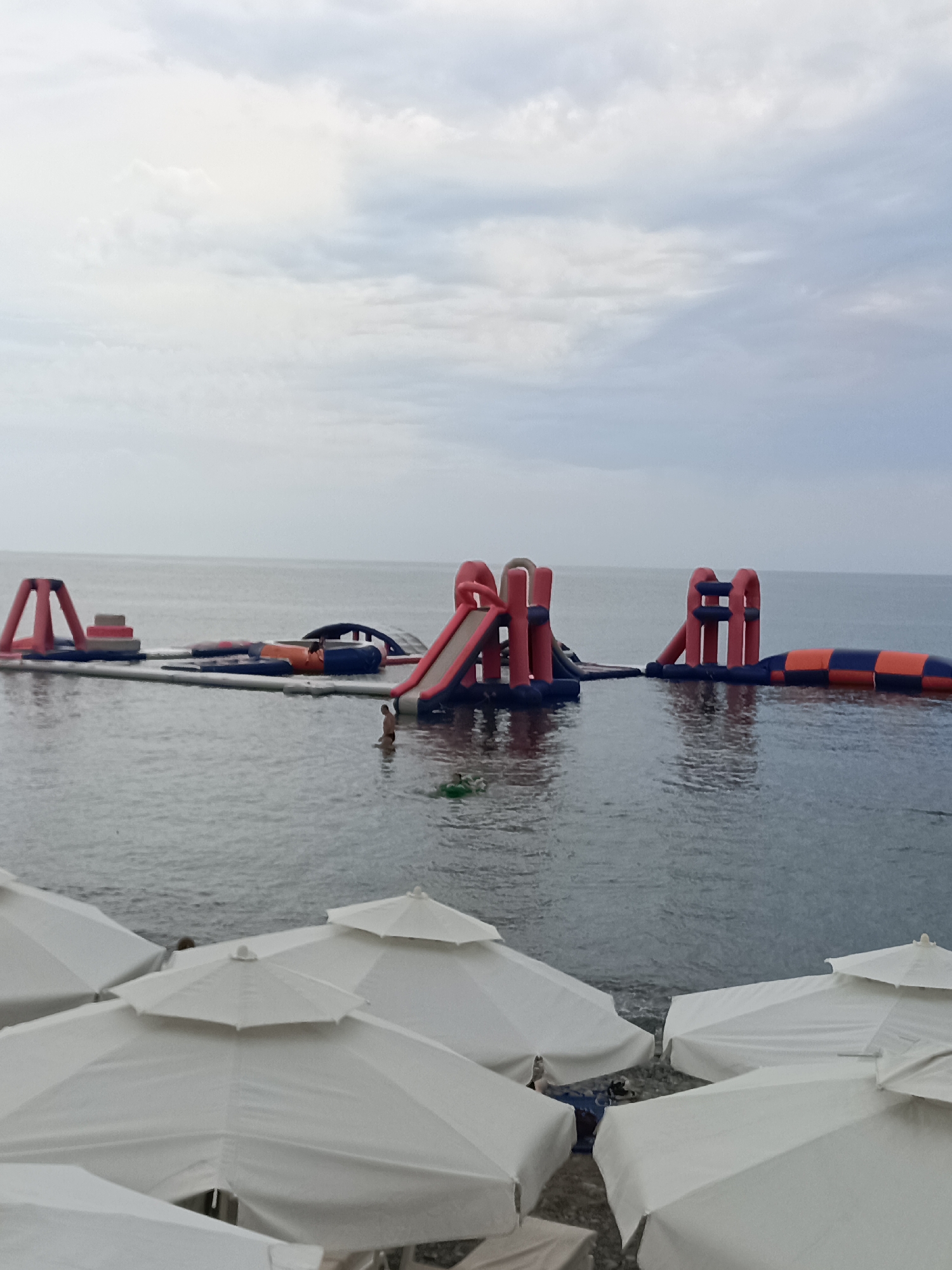 An aerial shot captures a vast expanse of the Black Sea off the coast of Sochi, Russia, under a cloudy sky. In the foreground, several white beach umbrellas are clustered, suggesting a leisure area. Further out, a vibrant inflatable water park is set up on the water, featuring red and blue structures, slides, and an obstacle course. One person is visible in the water near the inflatable park, appearing to swim. The sea is calm, reflecting the overcast sky and the colorful water park. The water is a muted blue-gray, with slight ripples. The horizon line is clearly defined where the sea meets the sky.