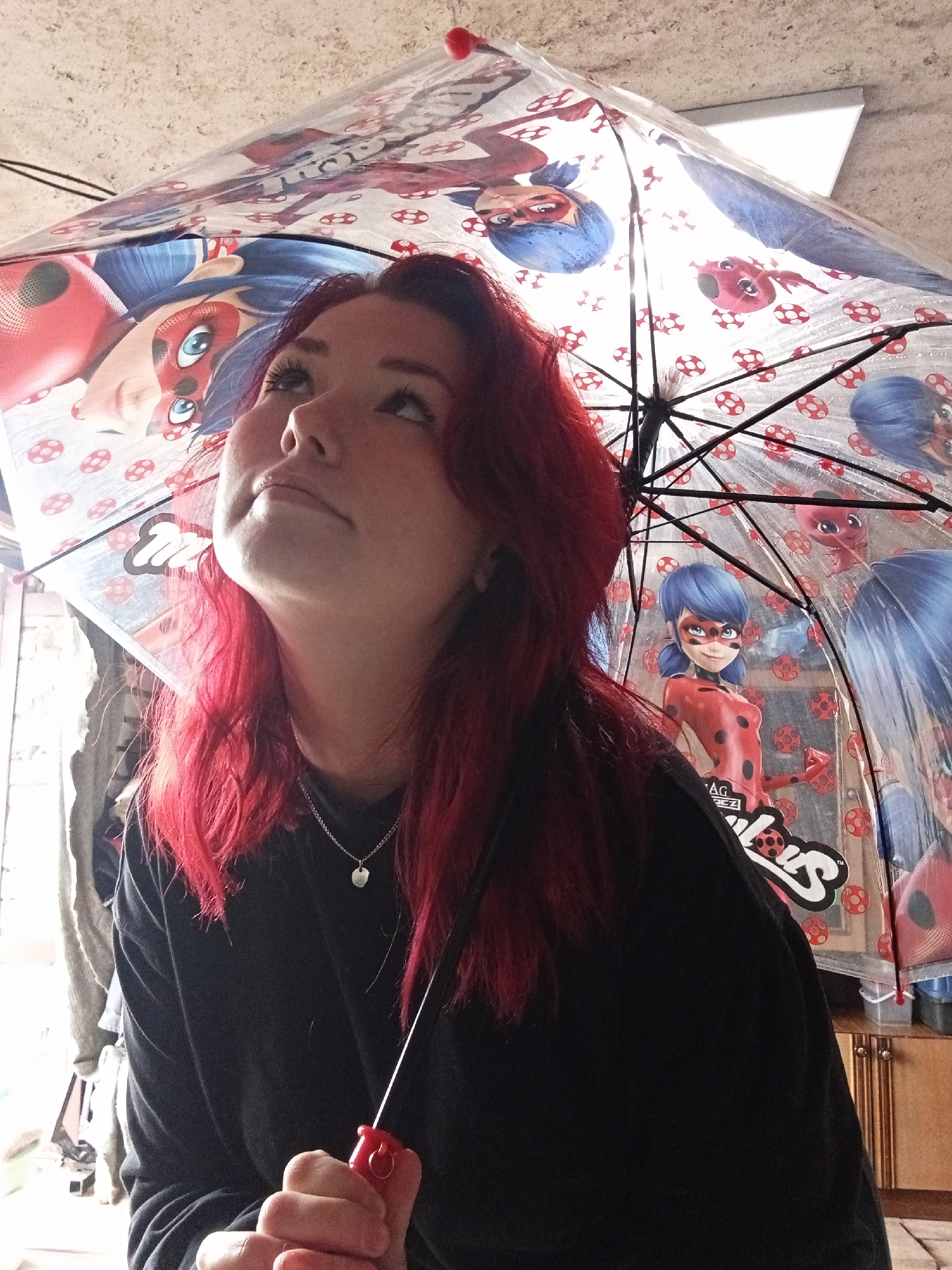 A young woman with vibrant red hair looks upwards under a clear umbrella adorned with characters from the animated series Miraculous Ladybug. The umbrella's translucent material allows light to filter through, illuminating the cartoon images and the woman's face. She is wearing a black long-sleeved shirt and a silver pendant necklace. Her left hand is visible, holding the red handle of the umbrella. The background suggests an indoor setting with a textured ceiling and what appears to be cabinetry to the right. The lighting indicates the presence of a light source from above or behind the umbrella.