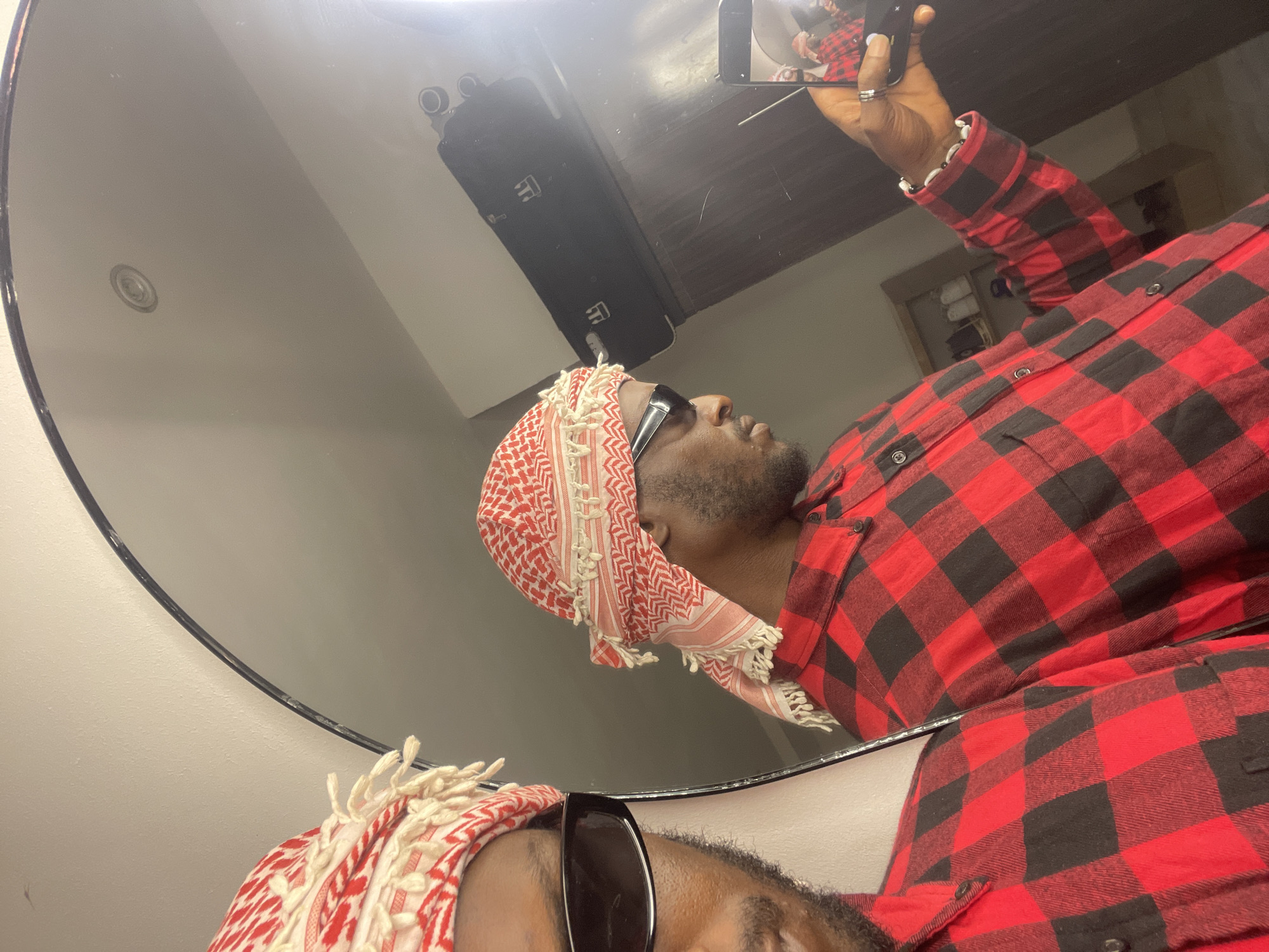 A young Black male is taking a selfie in a mirror, wearing a red and black checkered shirt and a red and white keffiyeh. He has dark sunglasses on. The mirror is large and oval-shaped, reflecting the man and the interior of what appears to be a bedroom or dressing area. In the reflection, there is a closet with some shelves visible, and on one shelf, a black bag with a white emblem. The man is holding a smartphone in his right hand, which is also reflected in the mirror, showing another image of him. The background is a neutral-colored wall. This scene is likely taking place in a residential setting within the city of Abuja, Nigeria.