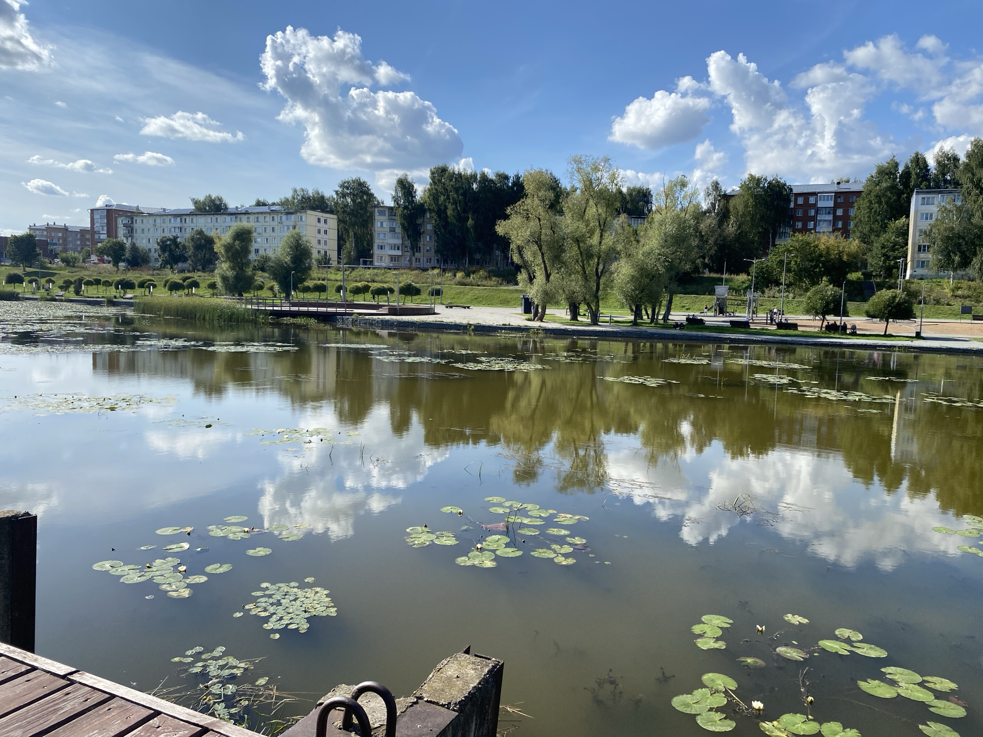 The image depicts a serene waterscape in Votkinsk, Russia, likely in a park or recreational area. A calm body of water, filled with lily pads and some blooming white water lilies, dominates the foreground. The water reflects the bright blue sky, dotted with fluffy white clouds, and the surrounding trees and buildings.
Along the far bank, a series of apartment buildings, characteristic of Soviet-era architecture, are visible, interspersed with lush green trees. A well-maintained pathway runs along the waterfront, with benches and lampposts suggesting a public space. Several people are casually strolling or sitting on the benches, enjoying the pleasant weather.
A wooden pier or dock juts out into the water on the left side of the frame, with concrete structures and metal railings visible at its edge. The overall impression is one of a peaceful, urban environment integrated with nature. The scene is captured on a clear, sunny day, with the sun casting gentle reflections on the water's surface.