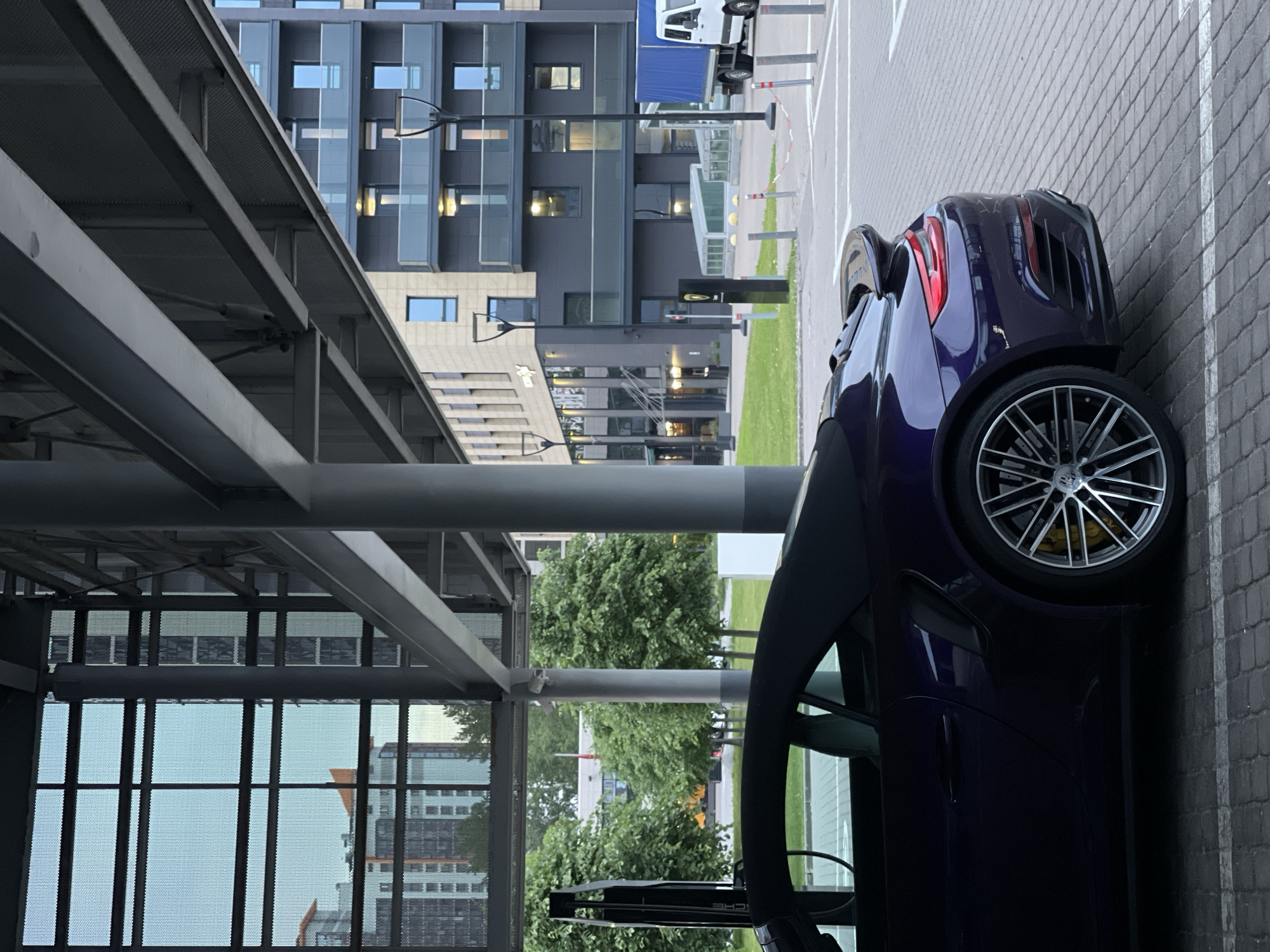 In an urban setting, a dark purple convertible Porsche is parked on a cobblestone surface under a modern overhang structure. The car's black soft top is up, and its rear spoiler is visible. The background reveals a contemporary building with multiple floors, featuring dark facades, balconies with glass railings, and numerous windows. To the left, through a grid-like facade, are glimpses of residential buildings. Further in the background, lush green trees line a grassy area, and a white truck with a blue tarp is parked near a lamppost. The scene suggests an outdoor parking area or driveway in front of a commercial or residential complex.