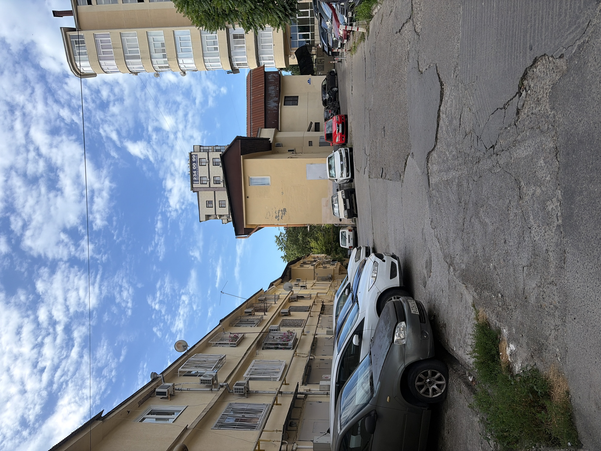 The image depicts a street scene in Sochi, Russia. In the foreground, a row of parked cars lines a cracked asphalt road. The cars include a dark gray sedan, a white hatchback, and a red sedan further down. Behind the cars, a long, pale yellow apartment building with numerous windows and air conditioning units stretches along the left side of the frame. Across the street, a multi-story building with a curved facade and many windows stands on the right. In the background, several other buildings of varying heights are visible, including one with a billboard displaying the number "234 00 99." The sky above is blue with scattered white clouds. Trees and greenery are visible on the right side of the image. The scene appears to be in a residential or urban area.