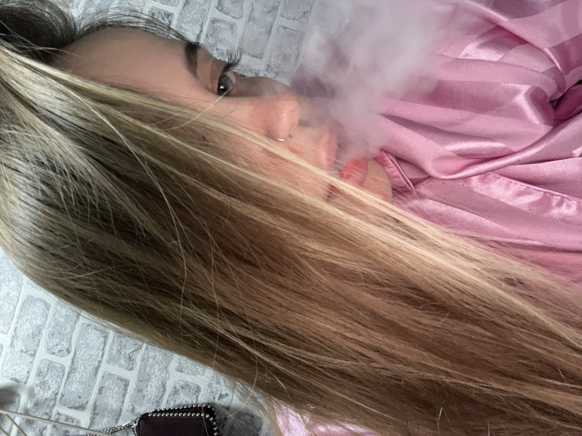 A person with long, light brown and blonde hair, a nose ring, and pink lipstick is lying down and exhaling a large cloud of white vapor. The person is wearing a pink striped satin top. The background features a white brick-patterned wall. A small, dark-colored purse with a silver chain is visible in the lower left corner. The location in the city of Novopokrovskaya, Russia is not evident from the image.