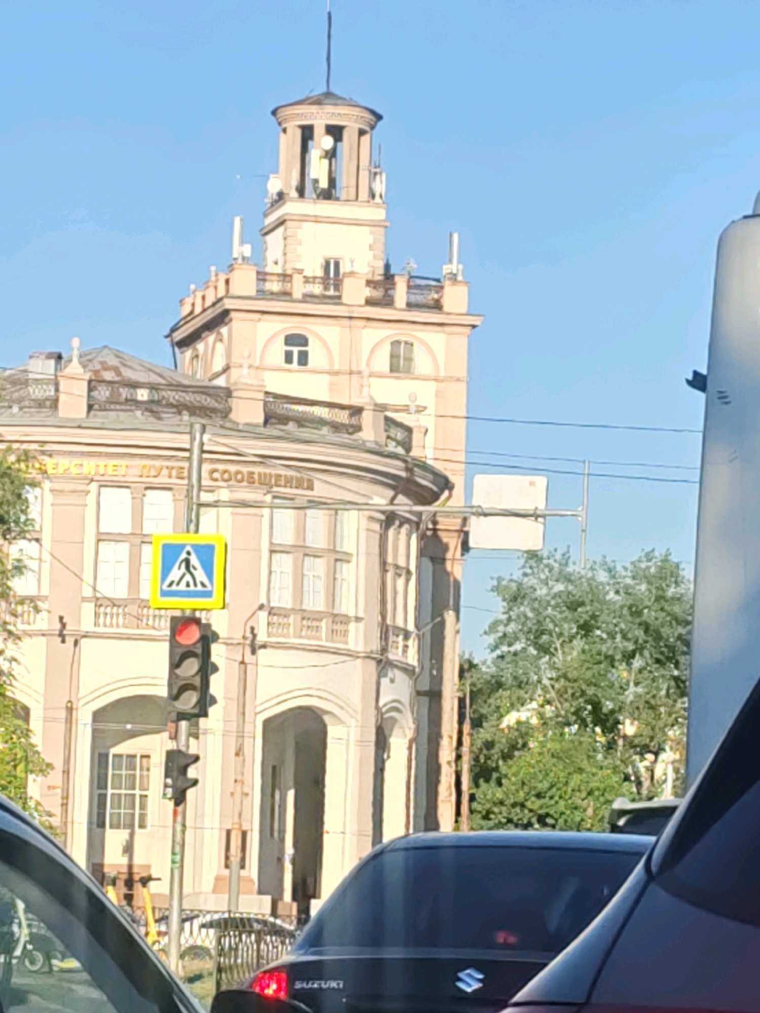 The image captures a street scene in Rostov-on-Don, Russia, dominated by a light-colored, multi-story building with a prominent tower. The building features arched doorways and windows, with decorative elements along its facade. In the foreground, a traffic light is visible, showing a red signal for vehicles and above it, a pedestrian crossing sign indicating a crosswalk. Several vehicles are present, including a dark SUV with "SUZUKI" branding on the rear and other cars partially visible. Green trees and foliage are interspersed within the urban landscape. The sky is clear and blue, suggesting a sunny day. The image appears to have been taken from within a vehicle.