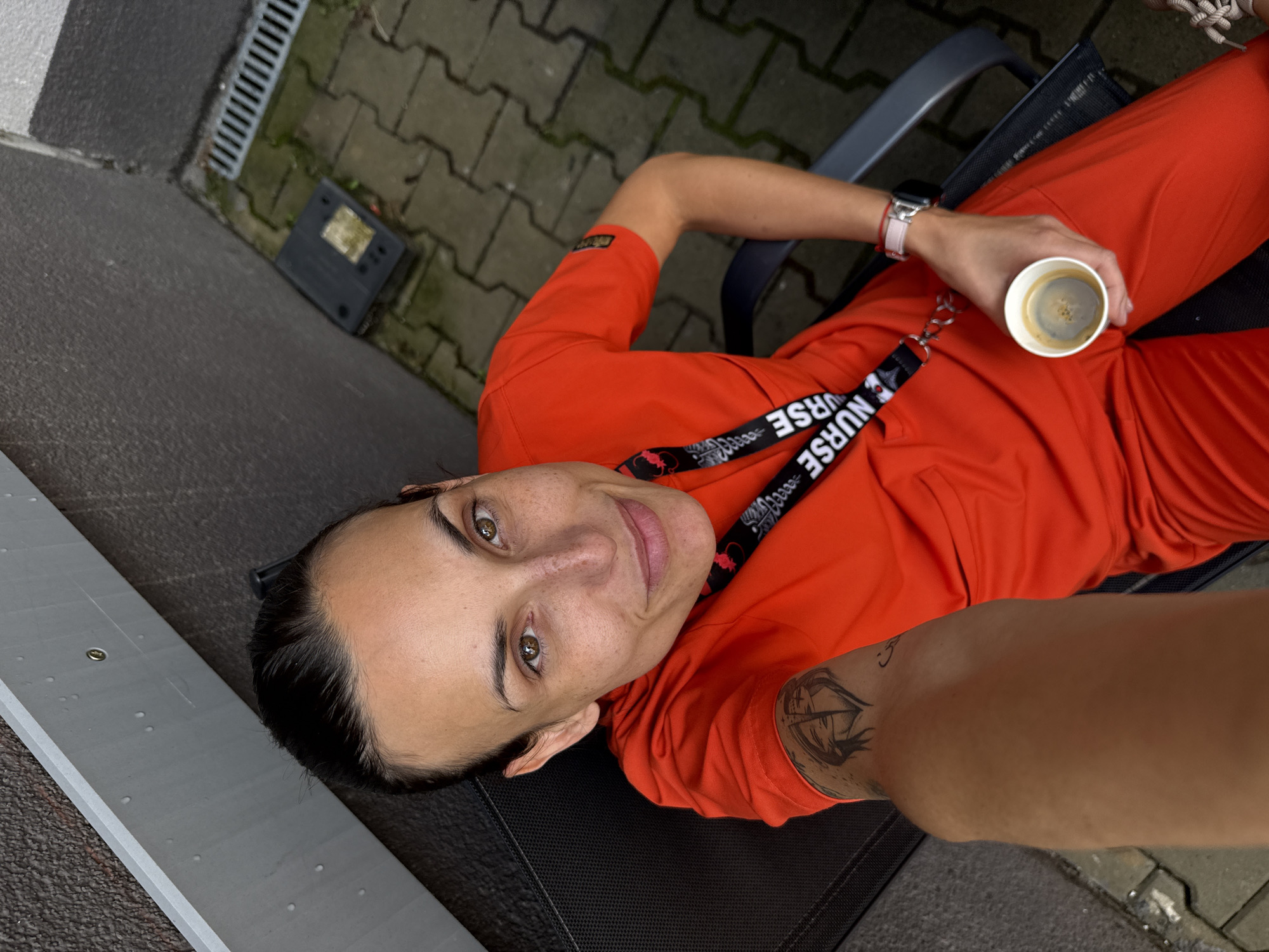 An overhead shot captures a woman, likely a healthcare professional, relaxing outdoors in Pleven, Bulgaria. She is lying on a dark mesh chair, wearing bright orange scrubs and a black lanyard. Her dark hair is pulled back, and she looks directly at the camera with a gentle smile, revealing brown eyes and light freckles.
In her right hand, she holds a white paper coffee cup containing a dark beverage. Her left wrist is adorned with a smartwatch featuring a pink band. A distinctive tattoo with an abstract design is visible on her right upper arm, near the elbow.
The background shows an outdoor setting with gray interlocking pavers, a metal drain, and a darker asphalt-like surface. A small, black, rectangular object with an illegible label rests on the ground near the drain. The lighting suggests it is daytime, possibly overcast, creating soft, even illumination.
The most prominent visible text is on her black lanyard, which clearly displays the word "NURSE" in white lettering, repeated along its length, along with some smaller, indistinct symbols. A small, dark patch with an unreadable logo is present on the right sleeve of her orange top. The overall scene depicts a moment of calm and relaxation for the individual.