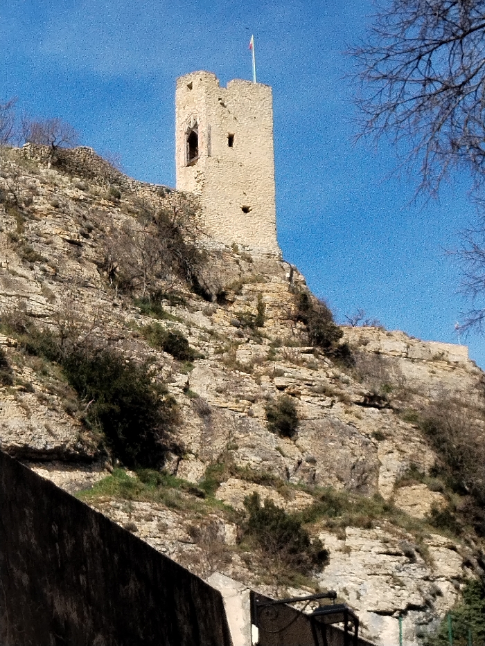 A stone tower with a small flag atop it stands prominently on a rocky hillside against a clear blue sky. The terrain is uneven, covered with sparse vegetation and layered rock formations. Bare branches of a tree are visible on the right. The base of a structure can be seen in the lower left, suggesting a human-made element at the bottom of the hill. The scene has a rustic, historical feel.