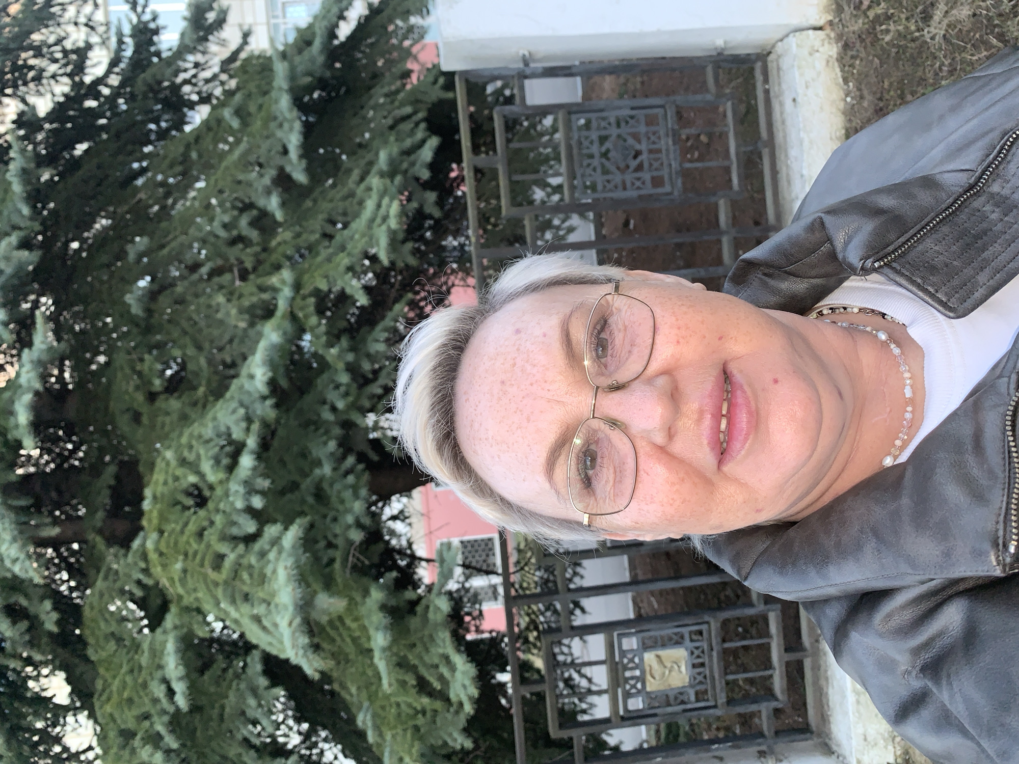 A fair-skinned, middle-aged or elderly woman with short, light blonde or grey hair is captured in a close-up, slightly off-center portrait. She wears glasses with thin gold frames and a white top layered under a dark, possibly black or dark brown, leather jacket with a zippered detail on the shoulder. A delicate beaded necklace, possibly with small pearls or light-colored beads, is visible around her neck, and a similar beaded bracelet adorns her wrist. She is looking towards the camera with a gentle smile, and freckles are noticeable on her face.
The background features a large, dense evergreen tree (likely a fir or spruce) with dark green needles on the left side of the frame. To the right, behind the woman, is a dark metal fence or gate featuring square decorative panels, some of which contain a lighter, possibly golden, emblem or pattern. Beyond the fence, parts of a building with light-colored walls and windows are visible. The scene appears to be outdoors during daylight hours, under soft light suggesting an overcast day or late afternoon. The setting is likely in Chaikovsky, Russia. The woman is posing for the photograph, and no specific activity is taking place. There is no visible text.