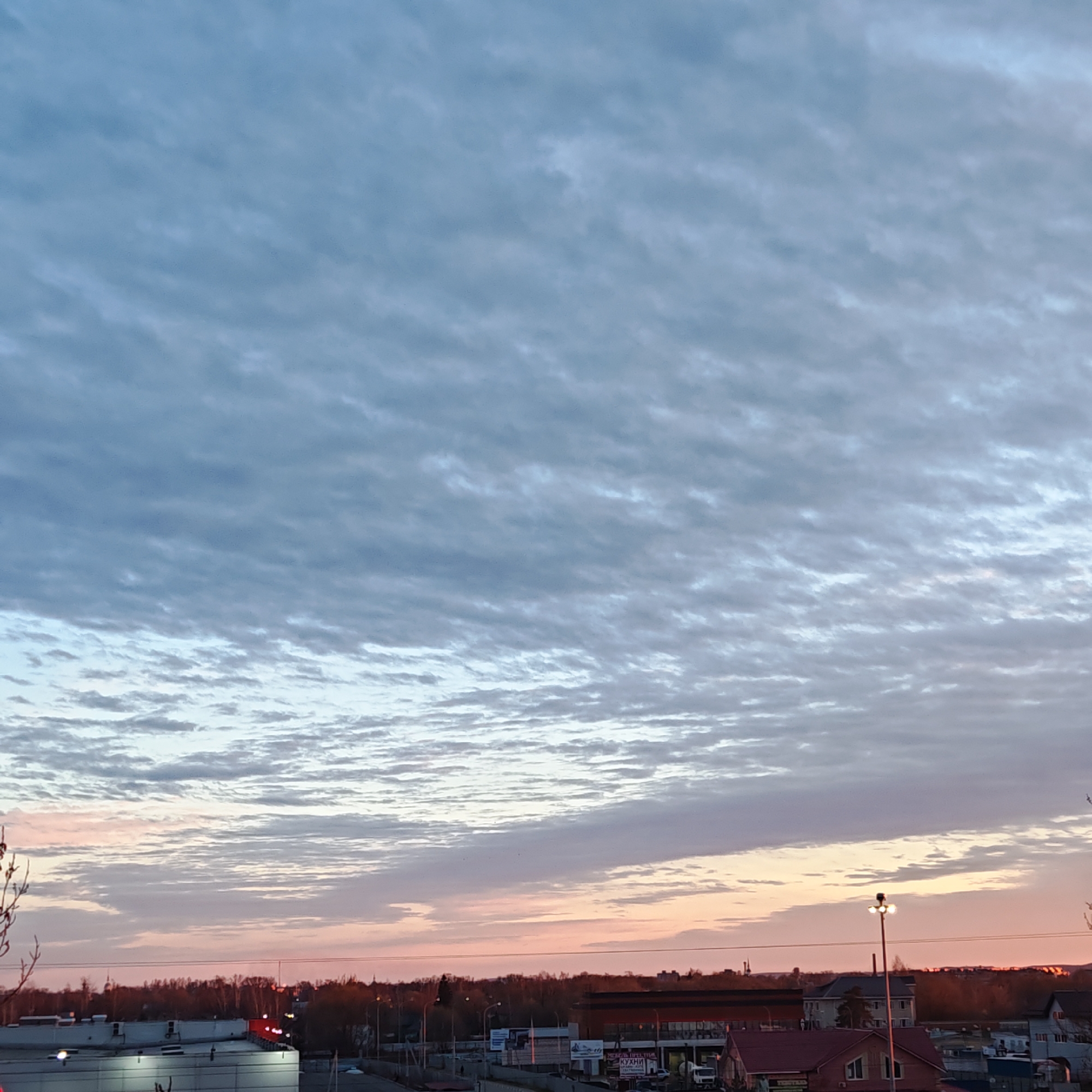 The image captures a cityscape under a sky filled with altocumulus clouds, indicating stable atmospheric conditions and potentially a change in weather. The sunset colors of pink and orange are visible near the horizon. The scene is likely in Rostov, Russia.
Foreground:
* A rooftop of a building, possibly a shopping center.
Midground:
* Trees form a line across the midground, separating the built-up areas from the sky.
* Buildings with varying roof colors (red and light-colored) are visible, with signs indicating commercial or residential zones.
* A street lamp stands out, illuminated against the twilight.
* A few cars are visible on the roads.
Background:
* The sky is the most prominent feature, with its textured altocumulus clouds.
* The color gradient from the setting sun adds depth and a sense of time.
Environment:
* The light suggests it is either sunrise or sunset.
* The trees suggest an urban setting with some greenery.