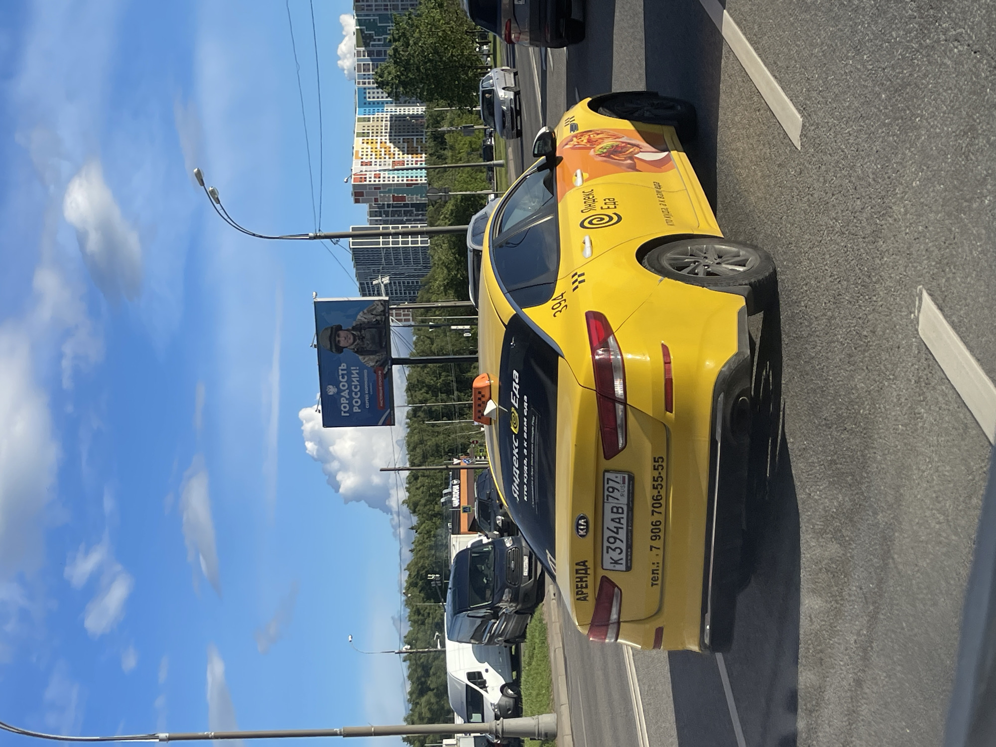 The image shows a yellow KIA sedan taxi with the "Yandex Eda" logo parked on the side of a road. The license plate number is K394AB 797. The phone number +7 906 706-55-55 is displayed on the car. A billboard featuring a military-themed advertisement ("Гордость России!") is visible in the background, along with some modern buildings. The location appears to be a street in Moscow, Russia. Other parked vehicles are seen on the roadside. The sky is blue with white clouds.