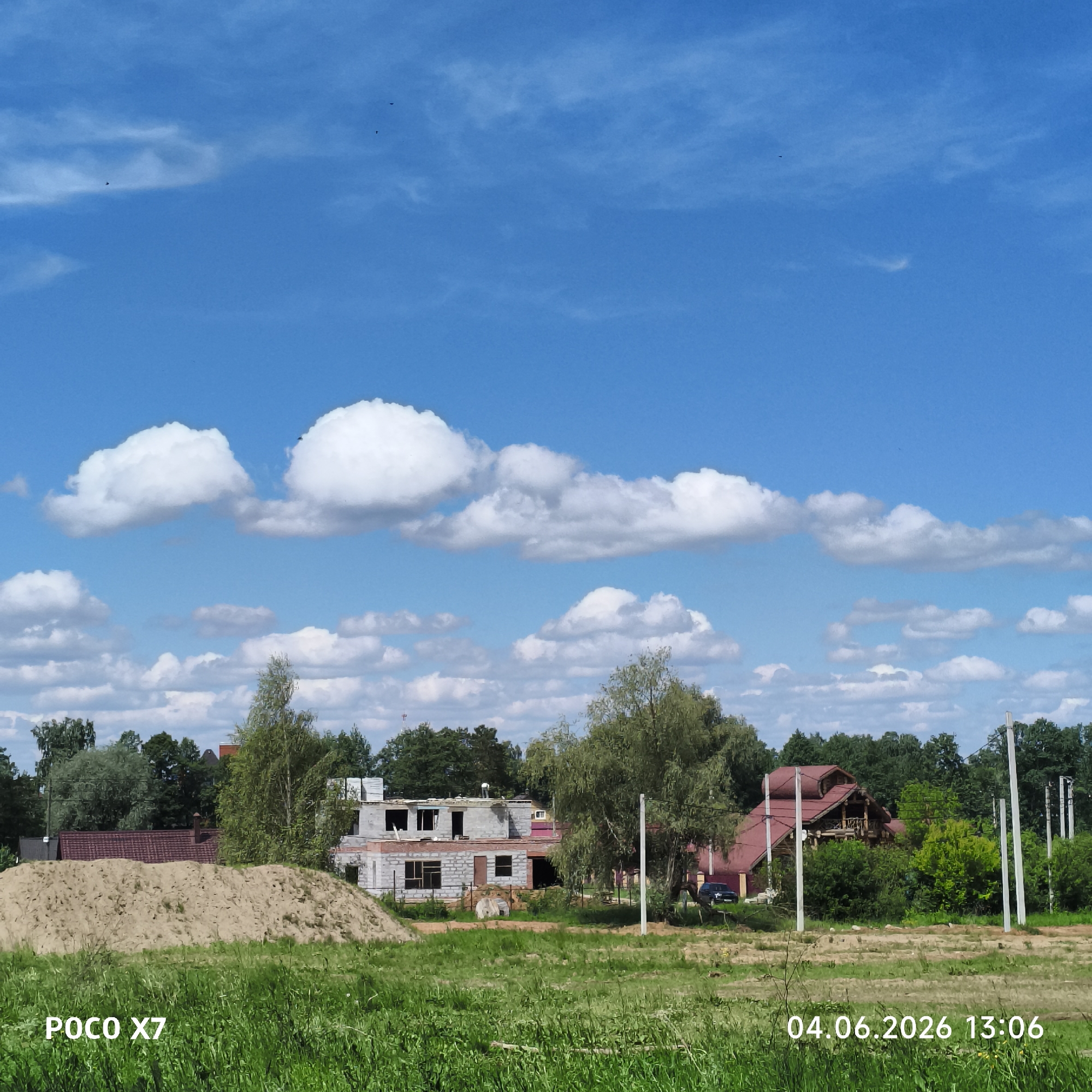 This image captures a vibrant, sunlit scene in a rural or developing suburban area of Chaikovsky, Russia. The setting is dominated by a bright blue sky filled with numerous fluffy white cumulus clouds, suggesting a clear and pleasant day.
In the foreground, a lush green field of grass stretches across the bottom of the frame, indicating healthy growth. To the left, a large, light-colored pile of sand signifies ongoing construction work. In the midground, a two-story house is under construction, its concrete block and brick walls partially erected with visible window and door openings. Behind this, another building with a reddish roof can be seen amidst trees.
Towards the right side of the midground and background, dense green trees form a natural barrier, with several prominent utility poles standing tall. Peeking through the trees on the right is a rustic-style house with a distinctive red-tiled, multi-peaked roof, and a dark-colored SUV or car is parked near it. Small, dark specks in the upper sky are birds in flight.
The time of day is indicated as early afternoon, 13:06, with bright sunlight illuminating the scene. The overall impression is one of development and natural beauty coexisting.
Visible text on the image includes "POCO X7" in the bottom left, likely indicating the device used to capture the photo, and "04.06.2026 13:06" in the bottom right, providing the date and time.