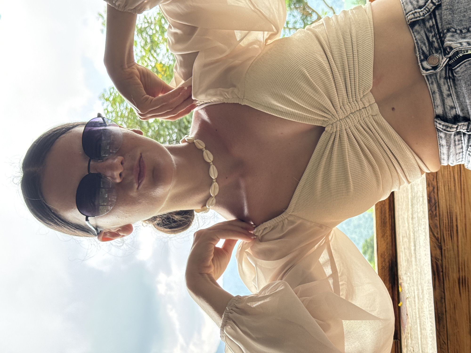 A young woman with dark hair pulled back and wearing round, dark sunglasses poses for a photo. She is dressed in a light-colored, ruched crop top with puff sleeves and a shell choker necklace. Her arms are raised, hands resting on her shoulders, and she is seated outdoors on a wooden surface. The background reveals a hazy sky and lush green foliage, suggesting an outdoor setting in a tropical or subtropical climate. The image appears to have been taken in Son Dien, Vietnam.