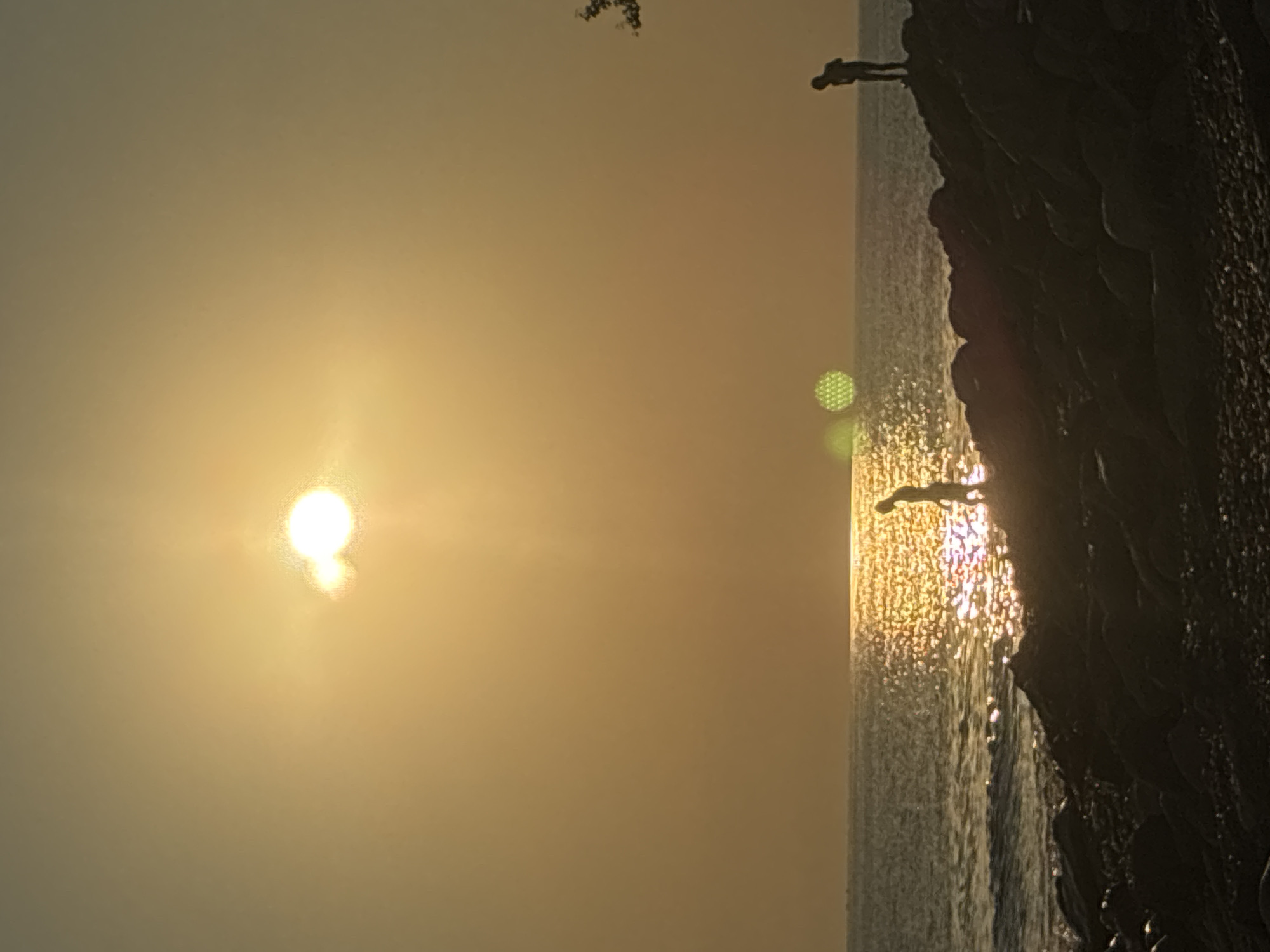 The image captures a sunset over the Black Sea in Sochi, Russia. The sun, a bright orb, is positioned in the upper left quadrant, casting a warm, golden light across the sky and water. The sea is calm, with gentle waves lapping against a rocky shore in the foreground. Two silhouetted figures are visible on the rocks. One person stands closer to the water's edge, looking out towards the sea, while the other is further back, also facing the water. The rocky shoreline is dark and textured, contrasting with the luminous water and sky. The overall atmosphere is serene and peaceful, with the fading light creating a sense of tranquility.