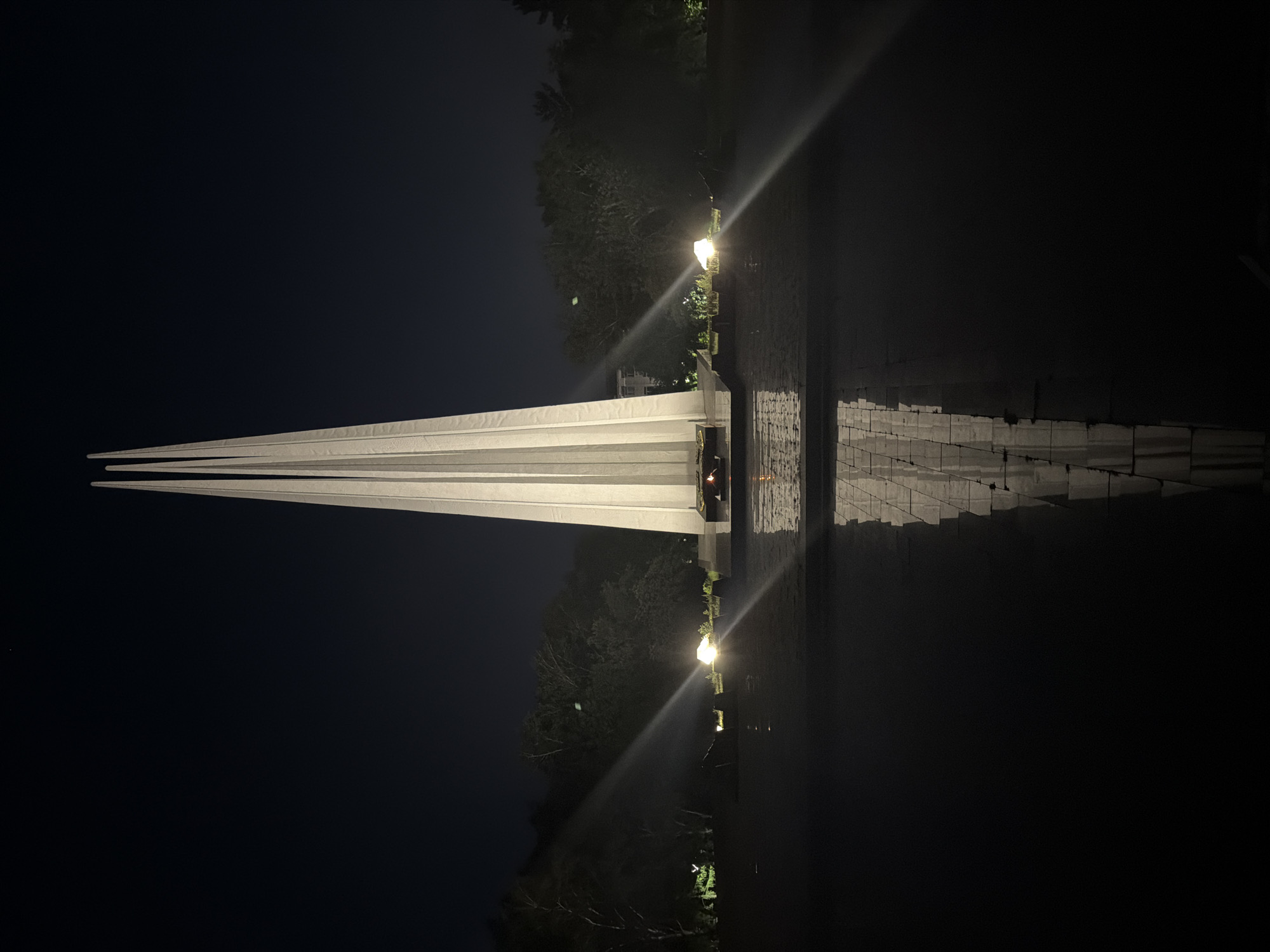 This image depicts the Eternal Flame monument in the city of Pavlodar, Kazakhstan, at night. The monument is a tall, white, obelisk-like structure composed of three slender, pointed sections that converge at the top. It stands in the center of a dark, reflective pool of water, which mirrors the monument. In the foreground, the water's surface is rippled, distorting the reflection slightly. Along the edges of the monument's base, there are small, burning flames, representing the eternal flame. The surrounding environment is dark, with trees visible in the background. Several spotlights are illuminating the monument and surrounding area, casting long beams of light that create a dramatic effect. The sky is dark and clear, with no visible stars or celestial bodies.