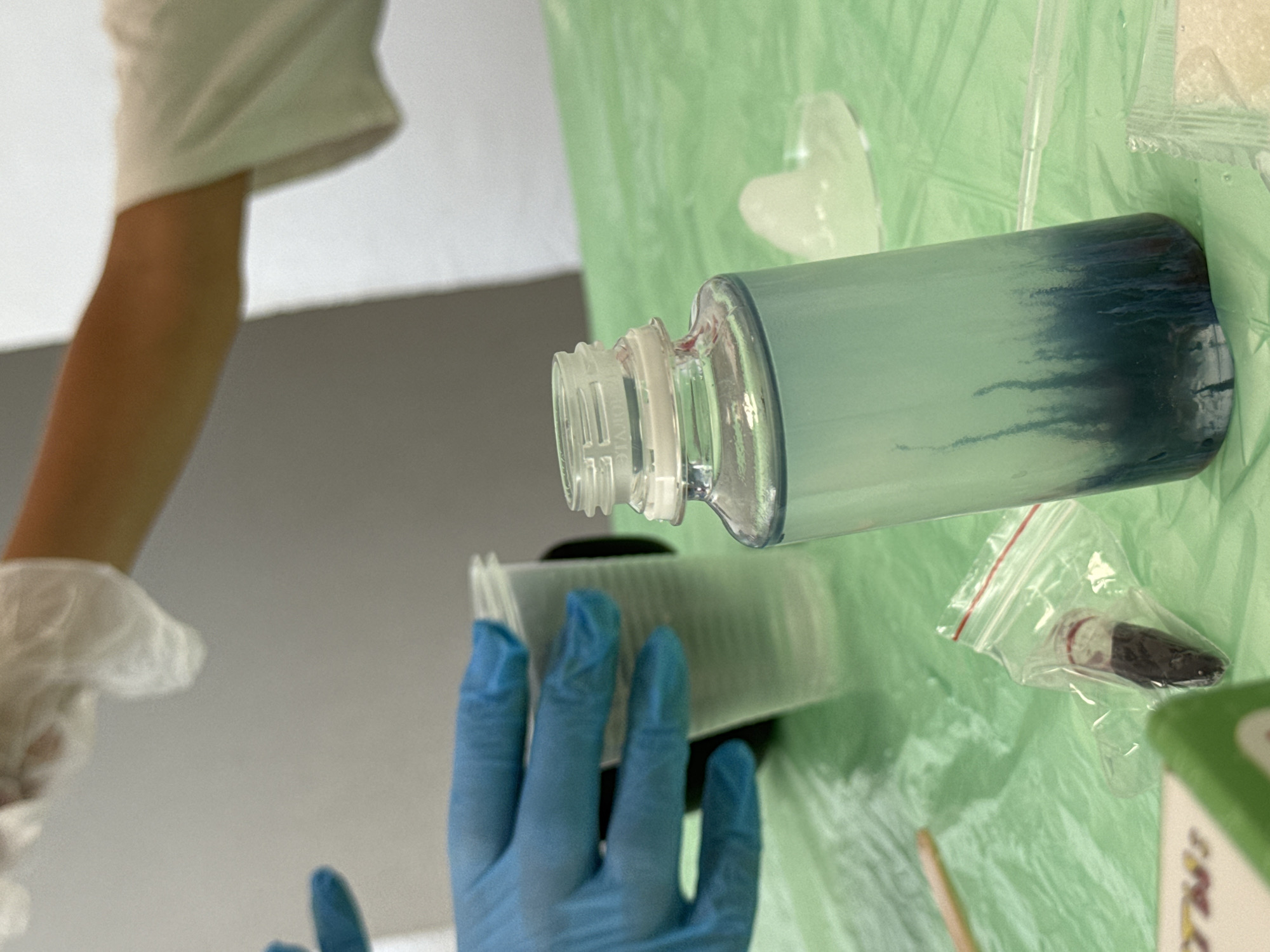 This is a close-up, eye-level shot of a science experiment on a green, crinkled plastic tablecloth, likely in Netanya, Israel, given the context. Two individuals are partially visible, both wearing protective gloves. The person on the left wears blue gloves and holds a clear plastic cup containing a substance. The person on the right, wearing white gloves, has their arm extended, suggesting they are either adding or preparing to add something to the experiment.
In the foreground, a clear glass bottle with a light blue liquid and darker blue streaks at the bottom stands upright. The bottle has a white plastic cap with a white sealing ring. To the left of the bottle, a small clear plastic bag contains a dark, viscous substance. A wooden stirrer lies diagonally on the tablecloth to the left of the bag. In the background, a white object, possibly a mold or another container, is visible, and a section of a wall or partition can be seen. The lighting suggests an indoor setting.