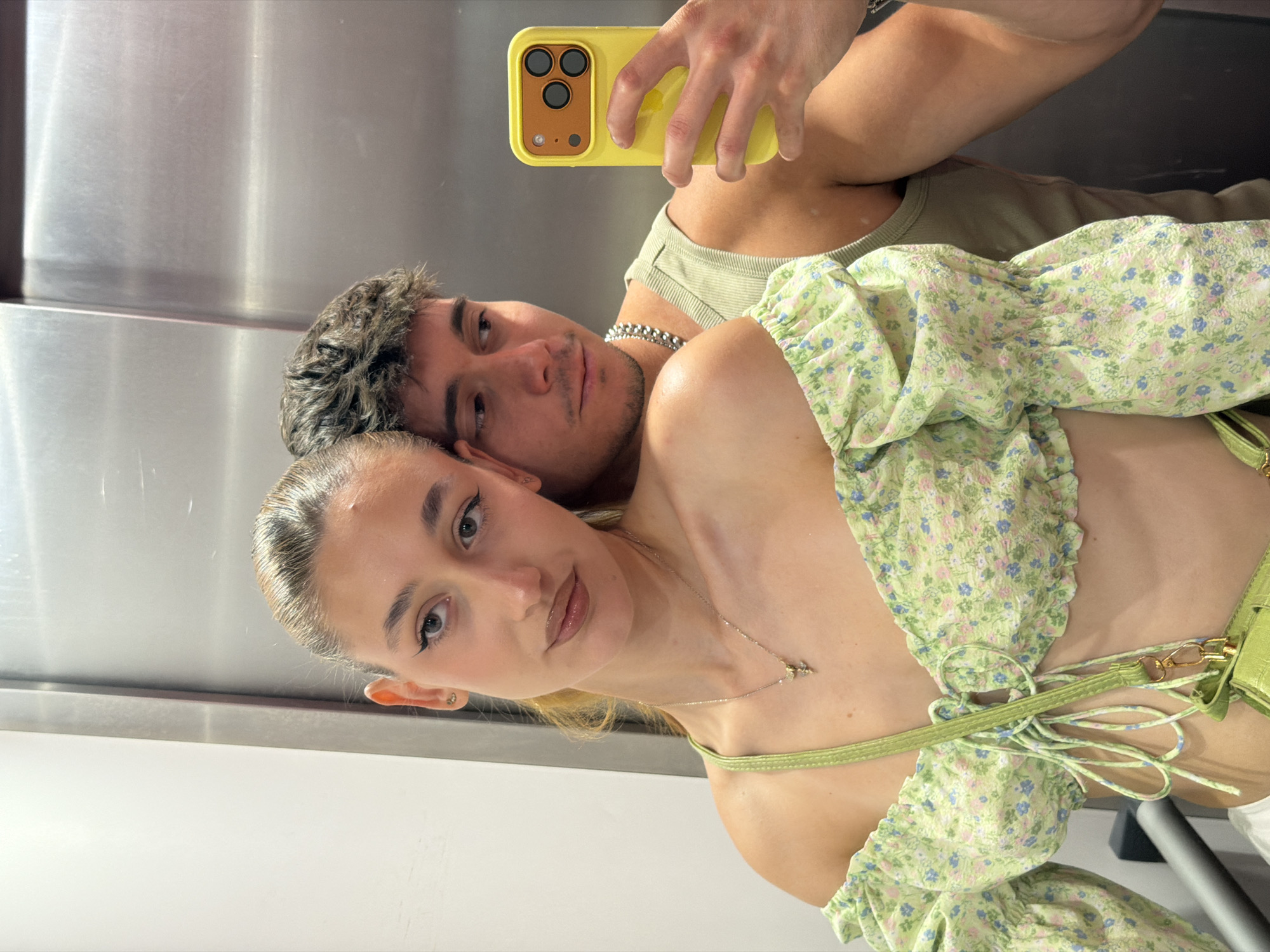 A young couple is taking a selfie in an elevator. The woman is on the left, facing the camera and resting her head on the man's shoulder. She has her hair pulled back, wears a light green off-the-shoulder floral top, and a silver necklace with a small pendant. The man is on the right, holding a yellow iPhone to take the photo. He wears a light green ribbed tank top and a silver chain necklace. The background of the elevator appears to be brushed metal. The scene is set in Sofia, Bulgaria, implied by the context of previous interactions.