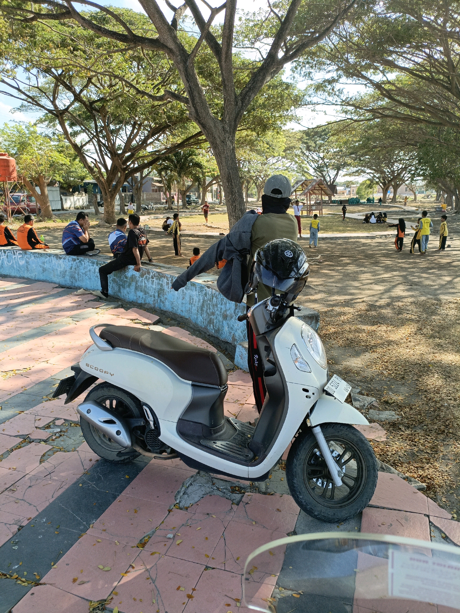A white and grey Honda Scoopy motorcycle is parked on a tiled pathway in a park area in Bulukumba, Indonesia. The pathway is bordered by a low concrete wall on one side, behind which people are sitting. In the background, several trees with dense canopies cast shadows on the ground. Children in yellow shirts are visible playing in the park, and a few adults are seen sitting or standing. A person wearing a green shirt and a grey cap is standing near the motorcycle, holding a grey jacket and a black helmet. The scene is well-lit by natural sunlight.