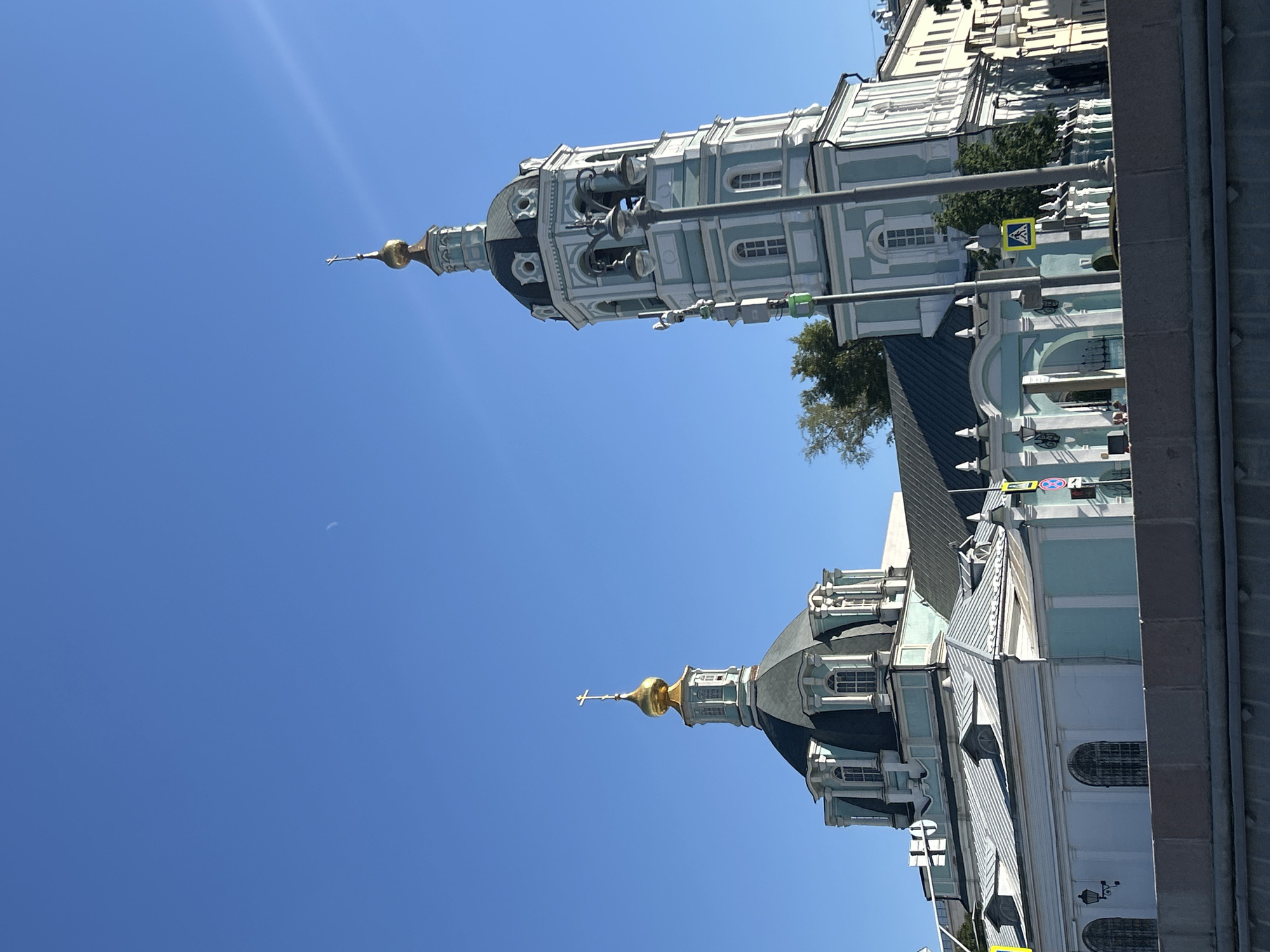 The image shows the Church of St. Clement in Moscow, Russia, a baroque style church with a tall bell tower. The church is painted white and light blue, with gold domes and crosses. The sky is a clear blue, with a thin crescent moon visible. The church is located on the Moskva River, with a stone embankment in the foreground. There are traffic lights and street signs visible, as well as some trees. The image is taken from a low angle, looking up at the church.