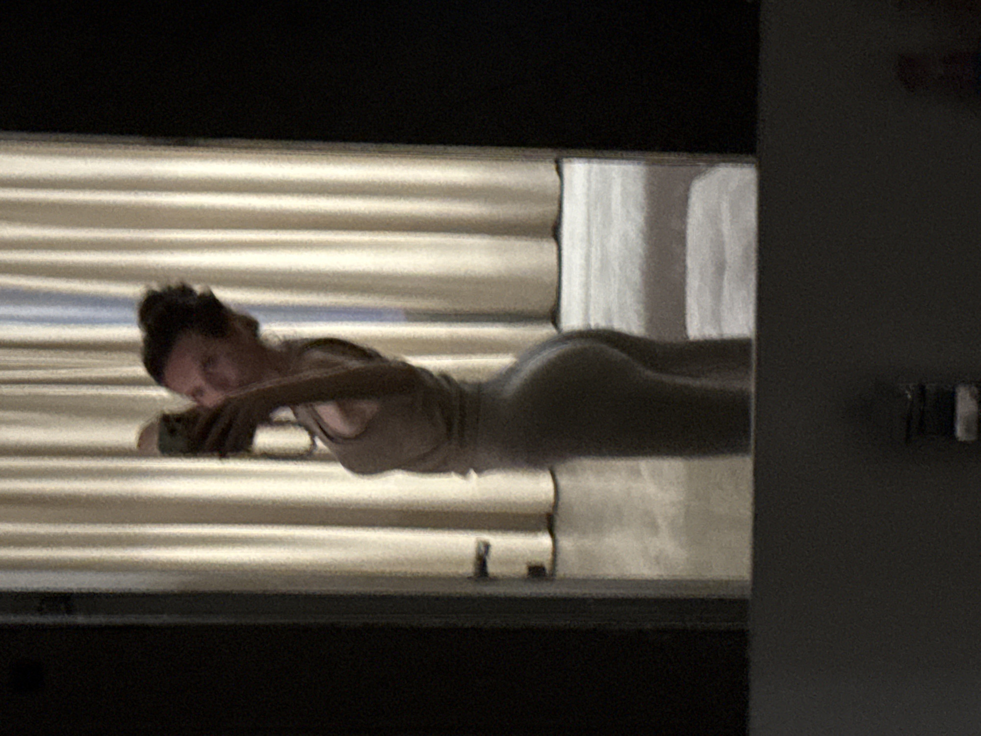 The image displays a person, likely a woman, lying prone with her body oriented horizontally across the frame. Her head is positioned towards the left, slightly lifted, as she looks down at a smartphone held in her hands. She appears to be wearing a light-colored top, possibly a tank top, and tight-fitting lower garments.
The background consists of horizontal blinds, which appear to be somewhat reflective or illuminated, creating a striped pattern behind the figure. To the far right of the frame, a dark, vertical structure, possibly a wall or piece of furniture, is visible. Below the person's torso, on the surface she is lying on, a few small, dark, indistinct objects can be vaguely discerned.
The overall lighting in the image is dim, suggesting it is either evening or nighttime indoors. The quality of the image is somewhat grainy, indicating low light conditions or a lower resolution capture. The activity depicted is one of quiet, personal interaction with a device. There is no visible text in the image itself. The location context provided is Khimki, Russia.