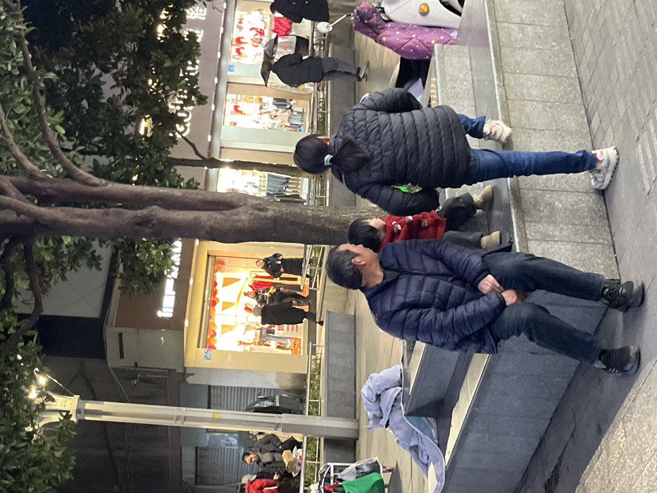 An evening street scene in Jiangjin, China, depicts a pedestrian area illuminated by artificial light from streetlights and commercial storefronts. In the immediate foreground, an adult male, wearing a dark blue puffer jacket, dark pants, and dark sneakers, is seated on a grey concrete barrier. To his right, a young child in a red jacket, dark pants, and light shoes sits beside him on the same barrier. Both individuals face towards the right side of the frame. An adult female stands to the right of the child, facing away from the camera, wearing a dark puffer jacket, blue jeans, and light sneakers, with her hair in a ponytail. A white scooter with a purple-patterned item draped over it is partially visible on the far right. A light blue garment is draped over the concrete barrier to the left of the seated man.
The midground features a paved walkway with several pedestrians. A tall streetlight with multiple lamps stands on the left. Several trees with sparse green foliage are present. Behind the pedestrians, multiple retail storefronts are brightly lit. The most prominent storefront displays clothing mannequins and features red decorative banners within its interior. Another storefront is visible to the right. Individuals in the midground are walking or standing, including one person holding a dark umbrella. The background consists of dimly lit building facades under a dark sky.