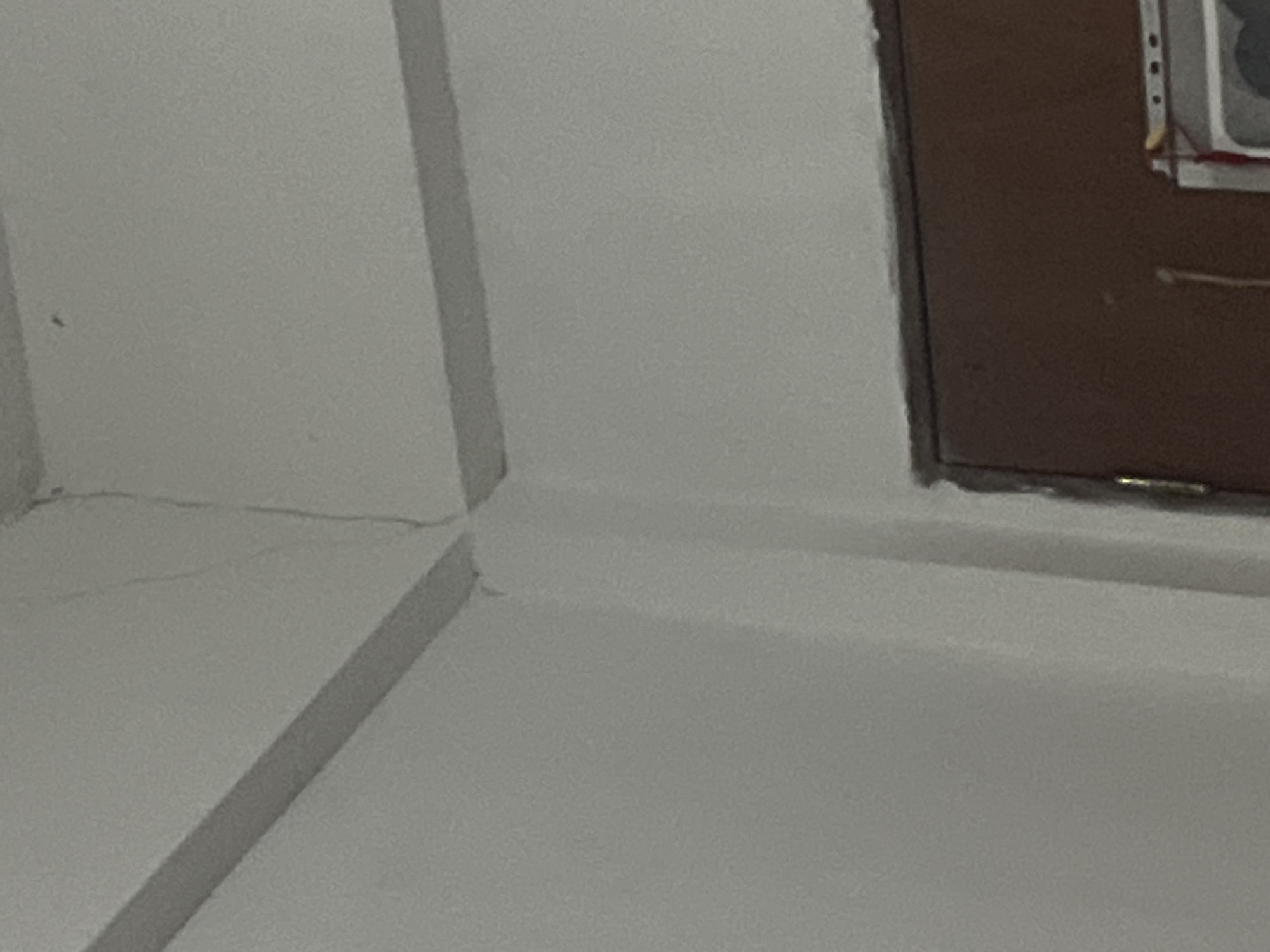 The image shows a close-up view of a white wall with a visible crack near the ceiling in a room in Santa Ana, Venezuela. The crack runs diagonally down the wall. There are also two horizontal moldings visible along the top of the wall. In the lower right portion of the image, a portion of a brown door is visible, with a small piece of what appears to be wiring or string hanging from it. The overall scene is indoor and appears to be a residential or commercial space.