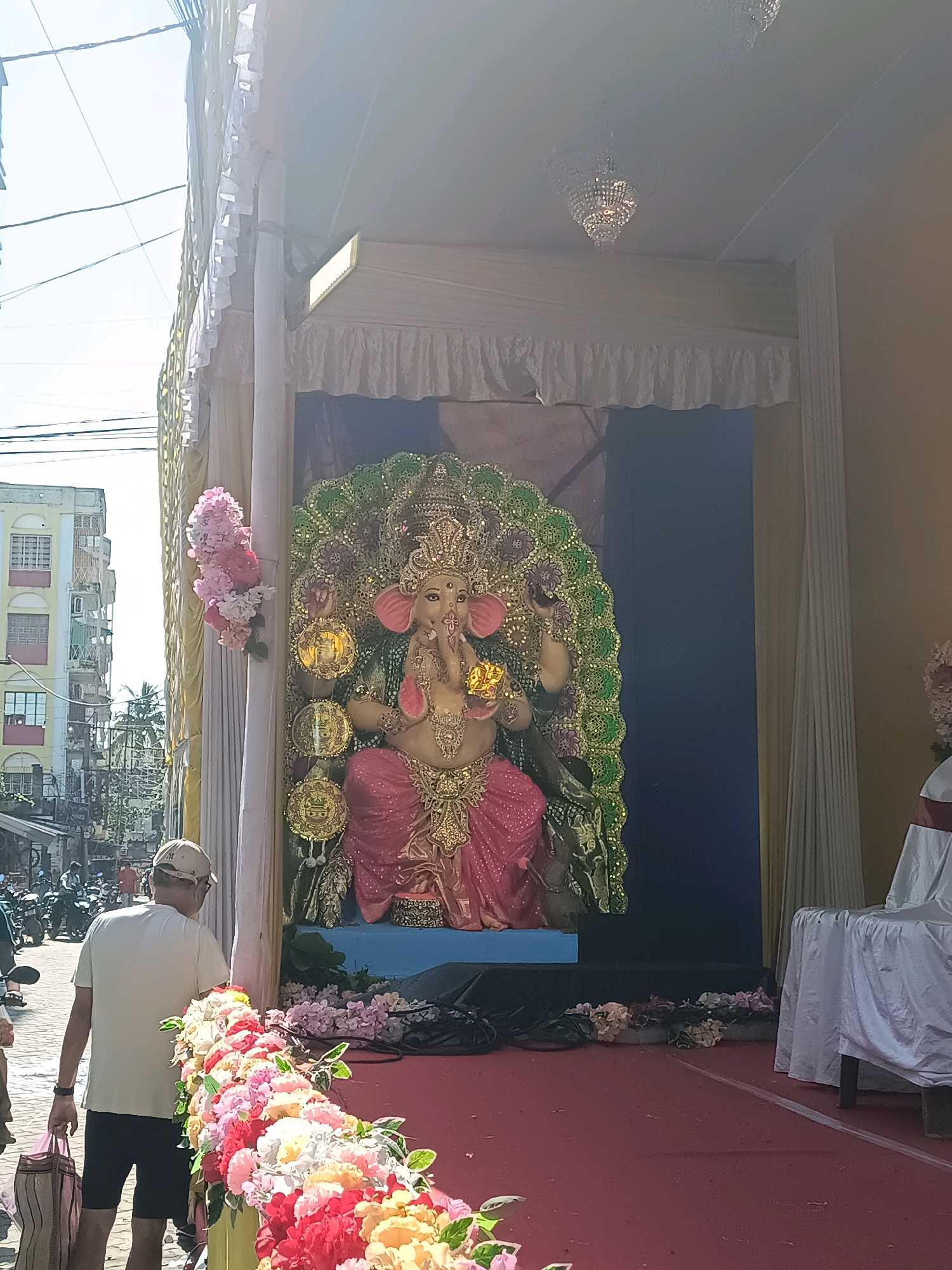 A statue of Ganesh is displayed outdoors in Panihati, India, under a tented structure. The statue is adorned with pink and gold clothing and a green, jeweled headdress. A man wearing a white t-shirt and a beige cap stands in front of the statue, carrying a bag. In the background, a building with yellow and white siding is visible. The scene appears to be part of a cultural festival or religious event.