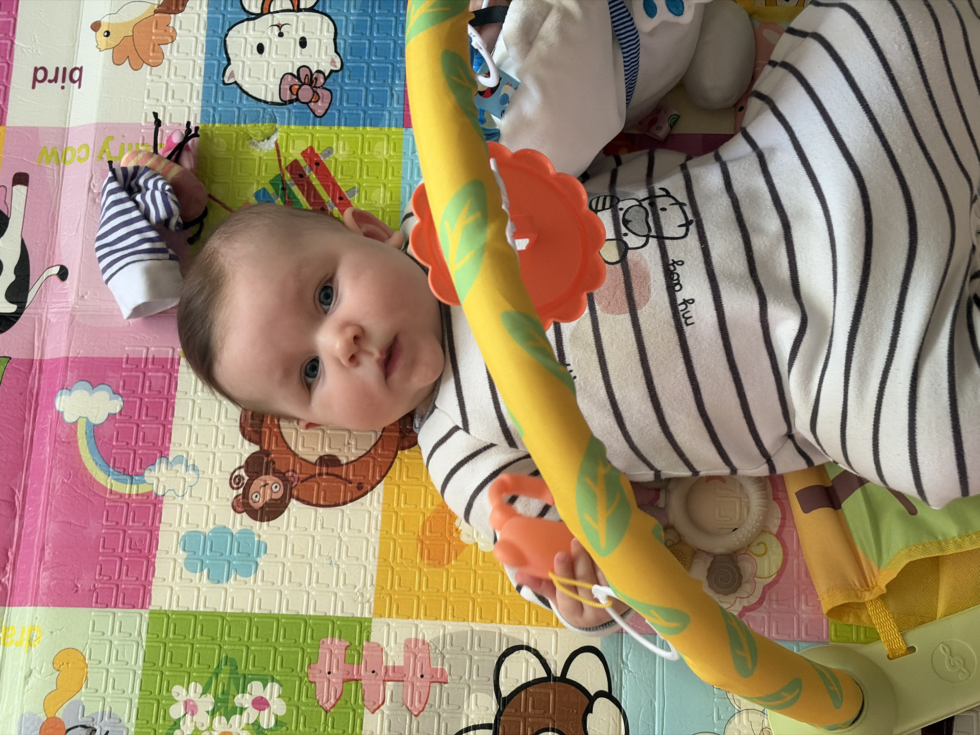 A baby wearing a striped onesie lies on a colorful playmat with animal illustrations. The baby is partially under a yellow and orange baby gym. A striped baby sock is nearby on the mat. The location is unknown, but the city of Chaikovsky, Russia, is mentioned.