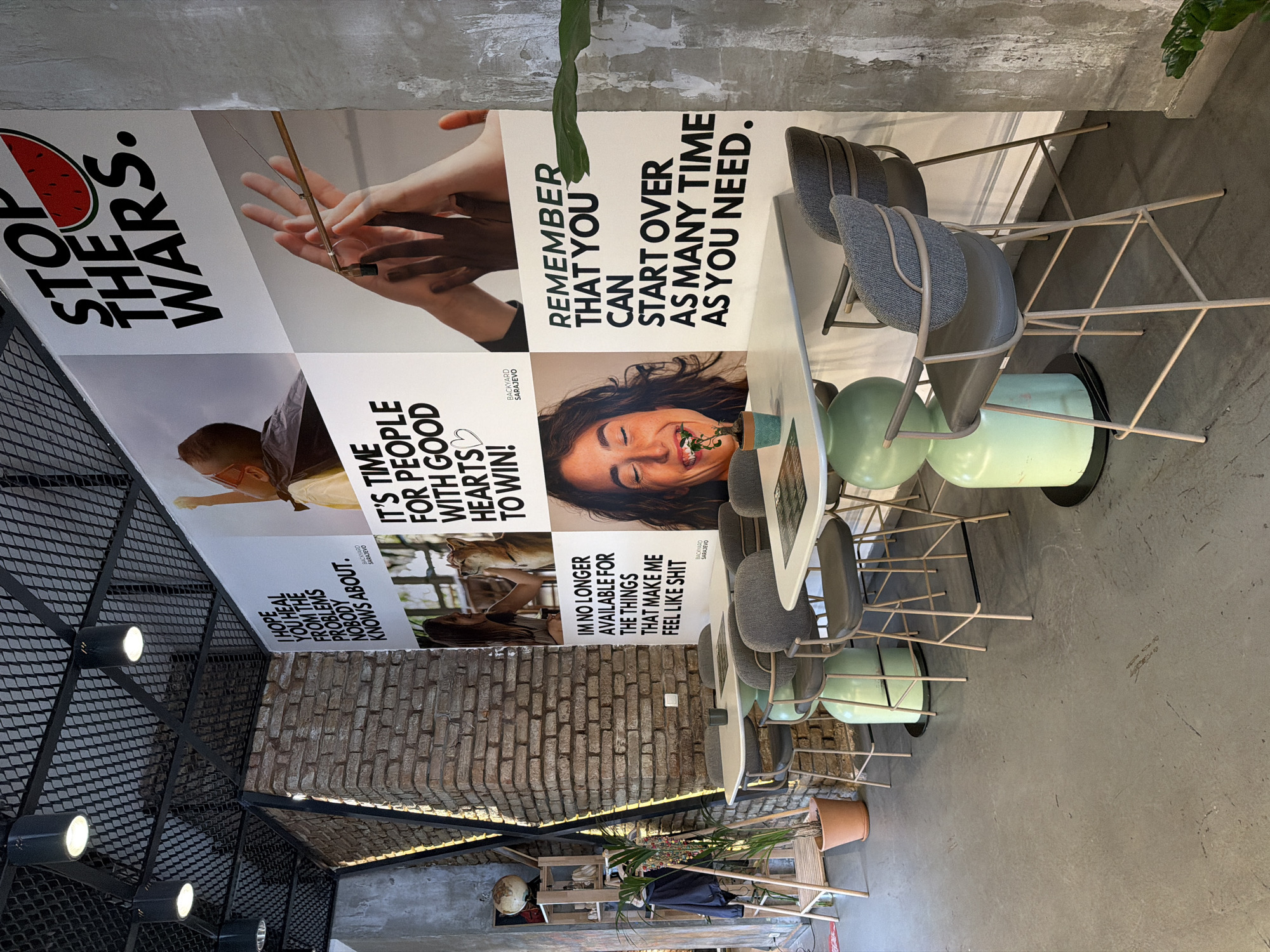 The image depicts the interior of a cafe with a prominent wall art featuring inspirational quotes and images. The wall art is divided into several panels. The top right panel has the text "STOP THE WARS." accompanied by a slice of watermelon. Below it, there's an image of hands reaching towards each other. To the left of this, a panel displays the text "I HOPE YOU HEAL FROM THE PROBLEMS NOBODY KNOWS ABOUT." Below that, a panel shows a woman with a dog and the text "IT'S TIME FOR PEOPLE WITH GOOD HEARTS TO WIN!". The bottom left panel features a woman with her eyes closed and a smile, with the text "IM NO LONGER AVAILABLE FOR THE THINGS THAT MAKE ME FEEL LIKE SHIT." The bottom right panel contains the text "REMEMBER THAT YOU CAN START OVER AS MANY TIME AS YOU NEED." The cafe has a modern industrial design with exposed brick walls, a metal grid ceiling with spotlights, and concrete floors. There are several high tables with bar stools, some with light green cylindrical bases. A plant is visible in a terracotta pot on the left side of the image, near a wooden shelf unit holding decorative items, including a globe. The overall atmosphere appears to be relaxed and contemporary. The presence of "BACKYARD SARAJEVO" on some of the panels suggests this establishment is located in Sarajevo, Bosnia and Herzegovina.