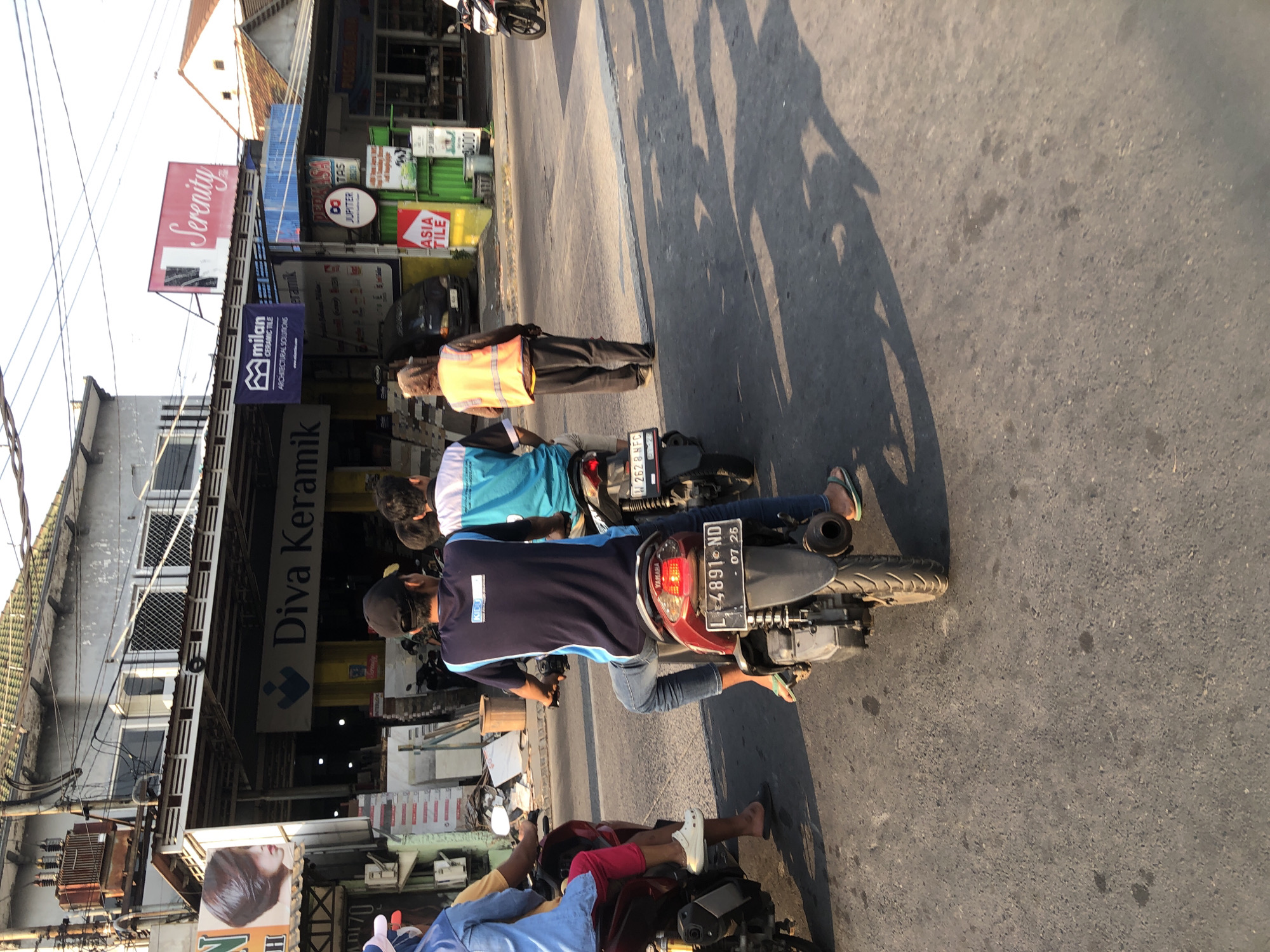 The image depicts a street scene in Boro, Indonesia, likely during daytime with visible sunlight casting shadows. In the foreground, three people are on motorcycles. The closest motorcycle has a rider wearing a dark blue shirt with a blue trim and blue jeans, a black baseball cap, and flip-flops. Behind him, another motorcycle carries two passengers. The person in front wears a light blue shirt, and the person behind wears a light blue and white patterned shirt. Further back, a person wearing a hooded jacket and an orange reflective vest is walking. The background features a row of buildings with awnings, displaying various signs including "Diva Keramik", "Serenity", "Milan Ceramic Tile", and "Asia Tile". There are parked motorcycles and a dark colored van visible in front of the buildings. The street surface is asphalt.