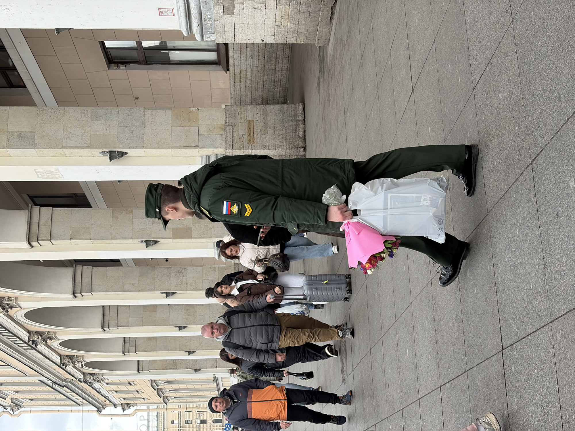 An outdoor scene captures several individuals on a paved urban sidewalk, set against a backdrop of a grand building with classical architecture, including arches, columns, and ornate stone facades. The central figure is a man in a dark green military uniform, complete with a cap and black polished boots. On his left sleeve, patches are visible: the Russian flag, a double-headed eagle emblem, and gold chevrons, indicating rank. He holds a vibrant bouquet of flowers wrapped in pink paper in his left hand, and a white shopping bag in his right.
Behind him, other people are present, some with luggage. A woman with a grey rolling suitcase stands near the military man. Further in the background, another woman is visible. In the foreground, an older man in a grey puffer jacket and another man in an orange and grey jacket are also standing on the sidewalk.
The setting appears to be a public square or a wide pedestrian area, typical of large European cities like Saint Petersburg, Russia. The building's beige and light-colored stone suggests a historic context. The lighting indicates it is daytime, possibly overcast, given the soft shadows. The overall impression is one of a public gathering or people waiting, perhaps at a transportation hub or a significant landmark. A small, unreadable red sign is high on the building, and faint large letters "OP" are visible on a building further in the distance.