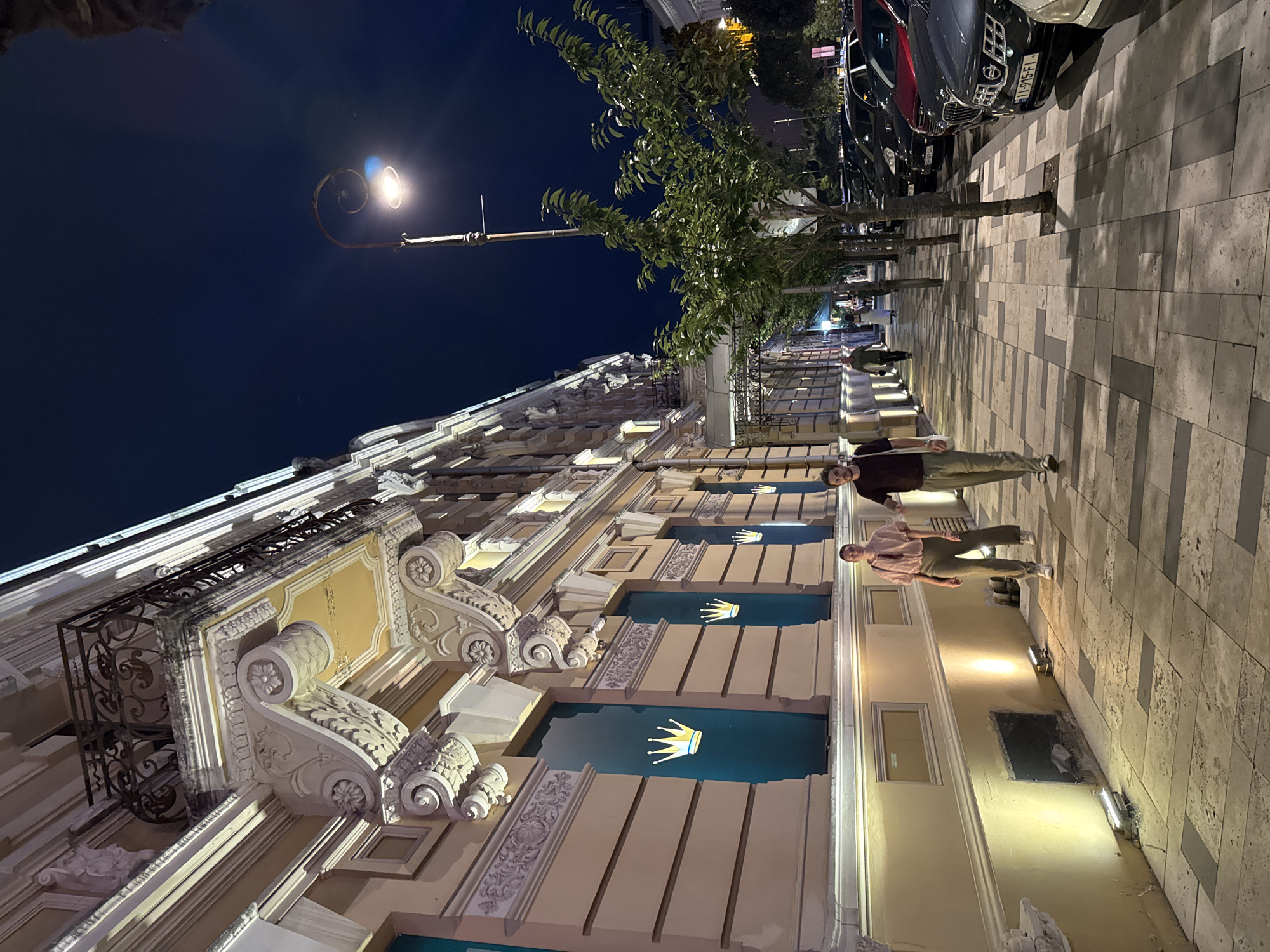 The image depicts a nighttime scene on a sidewalk in Batumi, Georgia. A man and a woman are walking on a paved sidewalk. The man, wearing a dark t-shirt and light-colored pants, carries a white bag over his shoulder. The woman, in a light pink shirt and similar light-colored pants, walks beside him.
To their left is a building with an ornate facade, illuminated by external lights. The building features balconies with decorative metal railings and elaborate architectural details. Several large windows on the ground floor display the Rolex crown logo on a teal background.
To their right, a line of parked cars is visible along the street. Streetlights illuminate the sidewalk and trees. The sky is dark, indicating it is night. The overall impression is of a well-lit urban street in Batumi.