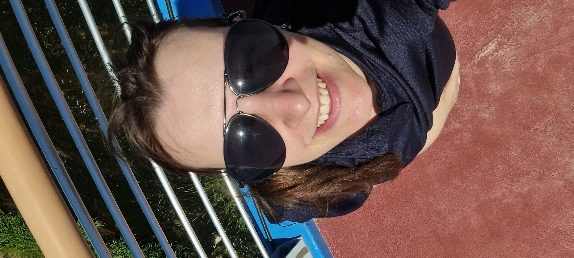 A smiling person wearing sunglasses stands on a bridge. The bridge has light brown supports and blue railing bases with steel guardrails. A stream flows under the bridge. The person is wearing a dark-colored shirt. The ground near the bridge is reddish-brown. The location is likely Dunnae-myeon, South Korea.