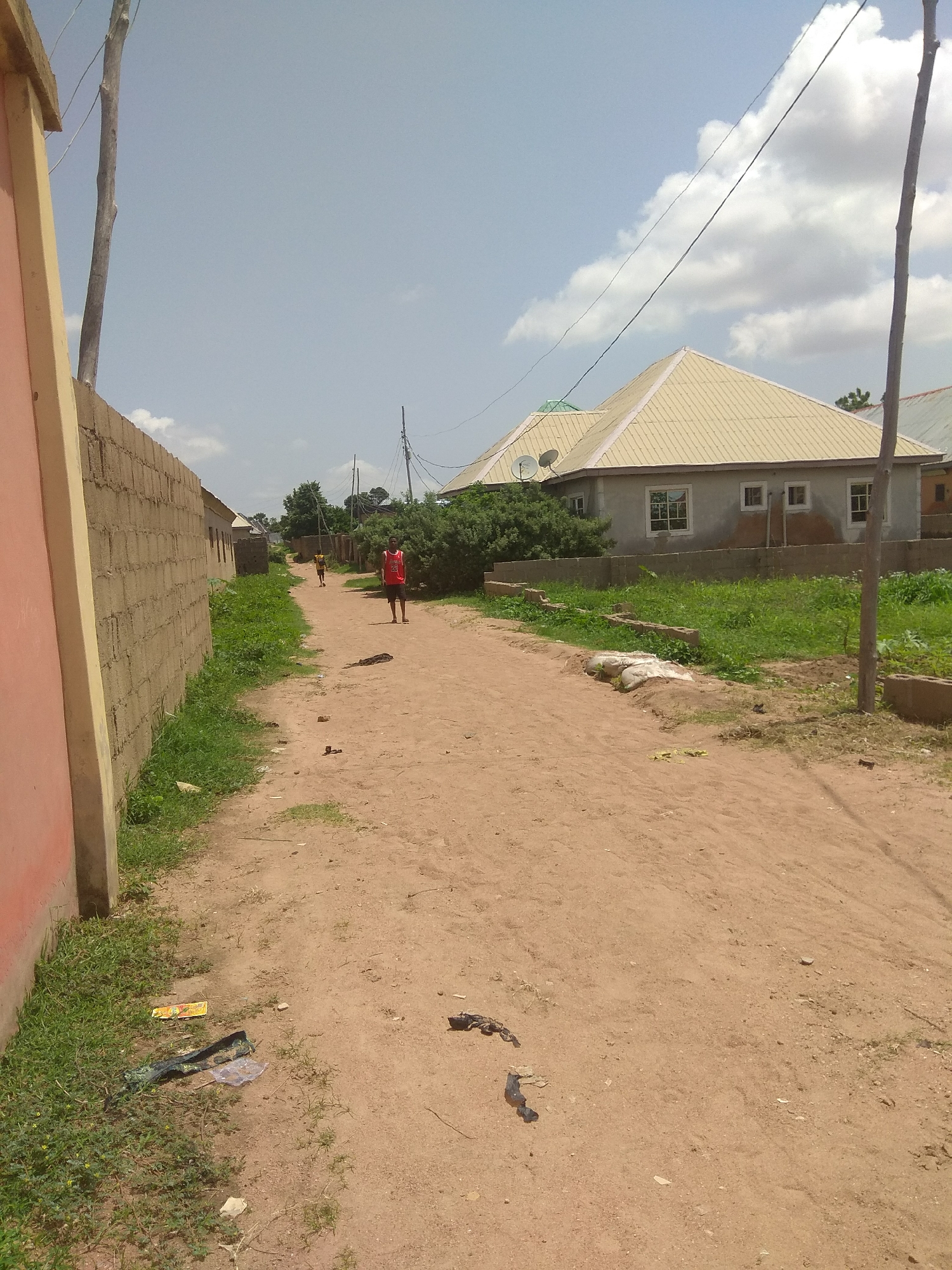 The image depicts a dusty, unpaved road in the city of Jalingo, Nigeria, lined with a low brick wall on the left and a residential building with a beige, corrugated metal roof on the right. The building features multiple windows and two satellite dishes mounted on the roof. Various trees and bushes are visible around the building and along the road.
Two individuals are present on the road. A young male wearing a red tank top with the number "23" on it stands near the center right of the frame, facing away from the camera and towards the distance. Further down the road, a smaller figure, possibly a child, is visible in the distance, wearing yellow clothing.
The ground is sandy and littered with debris, including plastic bags, wrappers, and a few scattered stones. A tall wooden pole with electrical wires running overhead stands on the right side of the road, parallel to the residential building. The sky is partly cloudy with scattered white clouds against a pale blue background. The scene suggests a casual outdoor setting in a residential area.