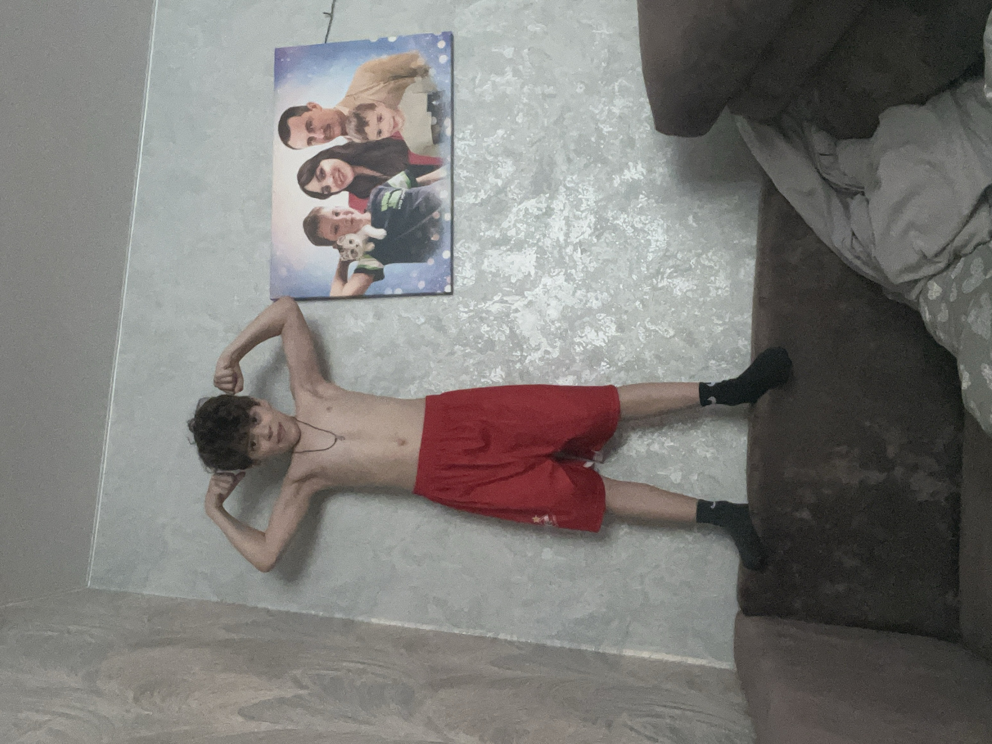 A teenager with dark, curly hair, shirtless, wearing red shorts and black socks, lies on a mottled gray surface, presumably a floor. He flexes his arms, facing the camera. Behind him, mounted on a pale gray wall, is a framed photograph of a family. To the right, a brown couch is partially visible, with gray blankets or cushions. The location is implied to be Noviy, Russia. No specific landmarks or details confirm this.