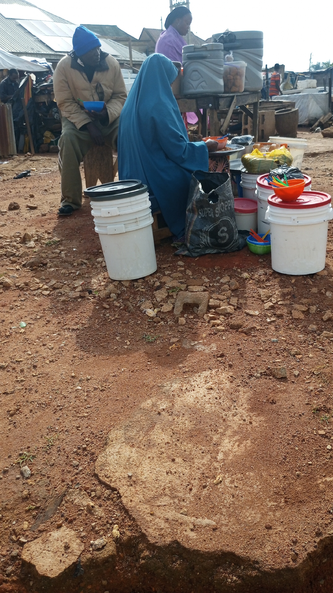 A medium shot captures an outdoor market scene in Gura Top, Nigeria. In the foreground, the reddish-brown earth is scattered with small rocks and debris. Two people are prominently featured. On the left, a man wearing a blue beanie and a tan jacket sits on a wooden stool, holding a small blue bowl. To his right, a person in a bright blue hijab, likely a woman, is partially visible, also seated. A black plastic bag with the words "SUPER SHOPPER" printed on it lies on the ground between them.
Behind the seated individuals, a wooden stall is laden with various items. Large grey plastic containers, white buckets with black lids, and red-lidded buckets are visible, some stacked. Brightly colored plastic utensils in green and orange bowls, along with yellow plastic bags filled with items, are also present on the stall. In the background, more market structures with corrugated metal roofs and scattered goods suggest a bustling commercial area. Another person, a woman in a purple top, is visible behind the stall, interacting with the items on display. The overall impression is one of a lively, if somewhat unkempt, local marketplace.