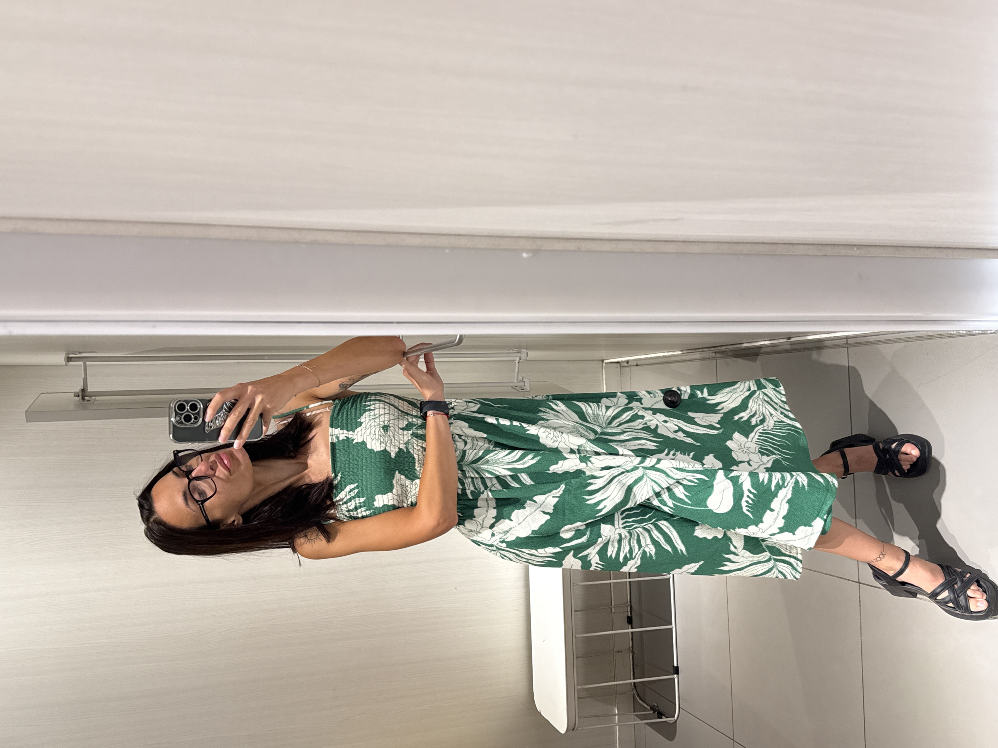 A woman is taking a selfie in a store fitting room. She is wearing a green and white floral print dress and black platform sandals. She has dark hair and is wearing glasses. She is standing in front of a mirror, and a white bench and rack are visible in the background. The floor is tiled. The fitting room appears to be in a store, possibly a clothing store, in the city of Pleven, Bulgaria.