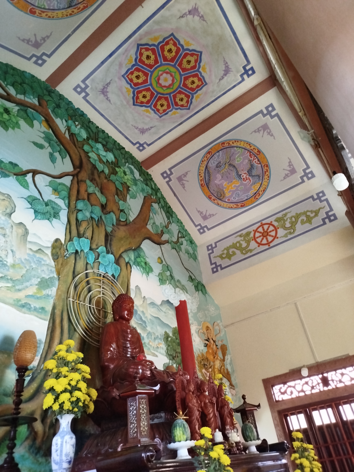 This media file captures the interior of a vibrant temple or pagoda in Liên Nghĩa, Vietnam, with a perspective looking upwards towards the altar and decorated ceiling.
At the center of the image is a large, reddish-brown statue of Buddha seated in a meditative posture, positioned on an ornate wooden altar. Behind the Buddha, a vivid mural covers the wall, depicting a grand tree with abundant green leaves, possibly a Bodhi tree, set against a backdrop of mountains and a blue sky. A white, circular fan-like grill is placed behind the Buddha's head, against the mural.
The altar in front of the Buddha statue is adorned with offerings and decorations. These include a vase of bright yellow chrysanthemum flowers, a tall red candle, and two watermelons (or similar fruit) placed on white stands. To the right of the main Buddha, several smaller, similarly colored statues are arranged on the altar. Further along the wall to the right, a painted mural of a multi-armed figure, possibly a Bodhisattva, is visible. On the far left, a tall, carved wooden stand holds a lantern-like object.
The ceiling is elaborately decorated with colorful traditional patterns and symbols. One prominent square panel features a circular mandala-like design with a central flower surrounded by red petals and intricate yellow details. Another square panel displays a circular motif of a dragon in purple and blue tones amidst clouds. A rectangular panel showcases the Dharma wheel (Dharmachakra) encircled by green swirling patterns. These decorated panels are framed by geometric blue and purple borders and separated by brown beams. A light fixture is visible on the ceiling towards the upper right.
The overall scene suggests a sacred space dedicated to worship and contemplation, with no specific activity taking place beyond the arrangement of devotional objects. The lighting appears to be a mix of natural and artificial light, indicating daytime. There is no visible text in the image. The architectural elements include light-colored walls and a wooden door or window frame with carved detailing on the right side.