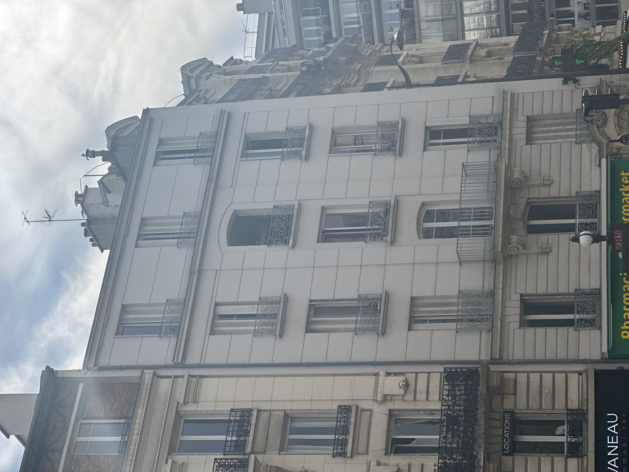 The image shows a street scene in Paris, France, focusing on the facades of several buildings. The architecture suggests a Haussmannian style, common in Paris. The central building is a light-colored residential building with multiple stories, each featuring windows with ornate iron balconies. The building to its left is an older structure with brick and stone detailing, also with balconies and windows. To the right, other buildings with varying architectural styles are visible, including one with a more modern appearance featuring many windows and balconies. The sky is cloudy, and the lighting suggests it is daytime. The ground level of the buildings appears to house commercial establishments, with visible signage, including "LOCATIONS," "VANEAU immobilier," and "Pharmacie." A street lamp and traffic signals are also present.