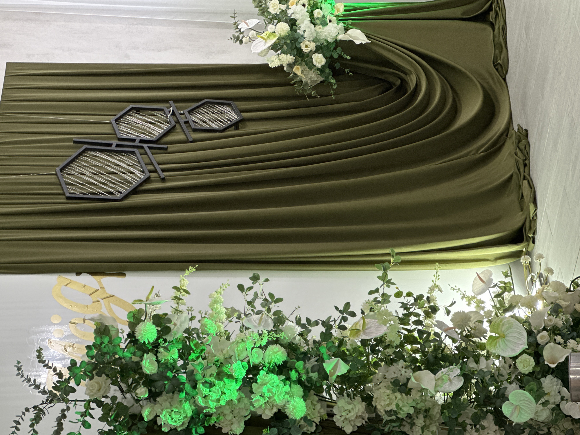 The image shows a decorative wall arrangement. A draped, olive green fabric serves as the backdrop, with its folds creating texture and depth. Two hexagonal decorative pieces, black-framed with what appears to be a beaded fill, are attached to the fabric. The bottom of the scene features lush arrangements of white and green flowers and foliage, some illuminated with a green light. The background behind the floral arrangement is white with a faint gold design visible.
