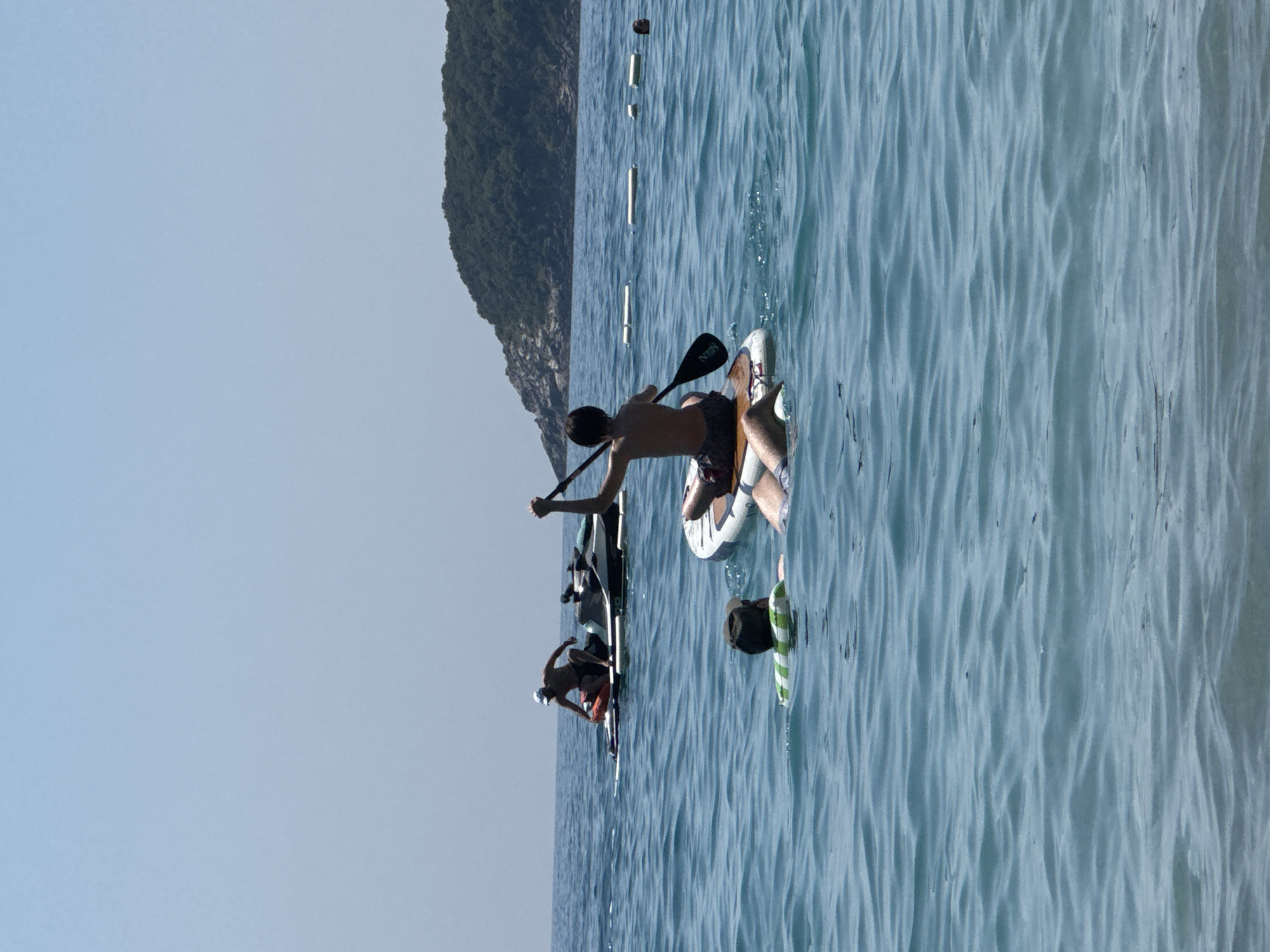 This vertical, medium-shot image captures a scene on the sea near Prijevor, Montenegro, on a clear, sunny day. In the foreground, two individuals are in the water. One person, wearing a green and white striped flotation device and a dark cap, is partially submerged. Another person is lying on a paddleboard, with their legs visible. In the mid-ground, another person is seated on a paddleboard, actively paddling with a black oar. Further back, two jet skis are visible. One jet ski is stationary on a paddleboard, with a person in a dark hat and shorts kneeling on it. Another jet ski is behind it. A wooded, hilly island with rocky outcrops forms the backdrop against a pale blue sky. White buoys are also visible in the water, creating a boundary.