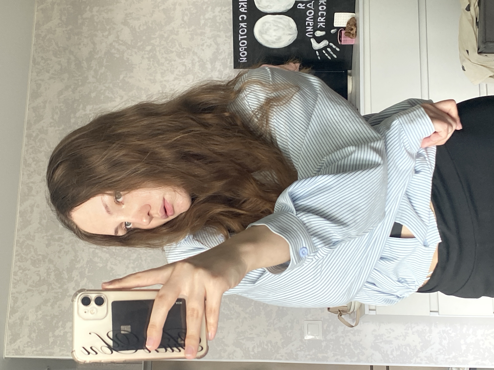 A young woman with long, wavy brown hair takes a selfie in a mirror. She is wearing a light blue and white striped button-down shirt, unbuttoned to reveal a black bra or camisole, and black pants. Her right hand holds a smartphone with a gold case and a black card insert. The background appears to be an indoor setting, possibly a bedroom, with a textured, light-colored wall. To the right, a black board with white drawings and text is visible on a white dresser. The scene suggests a casual moment, perhaps at home in Izhevsk, Russia.