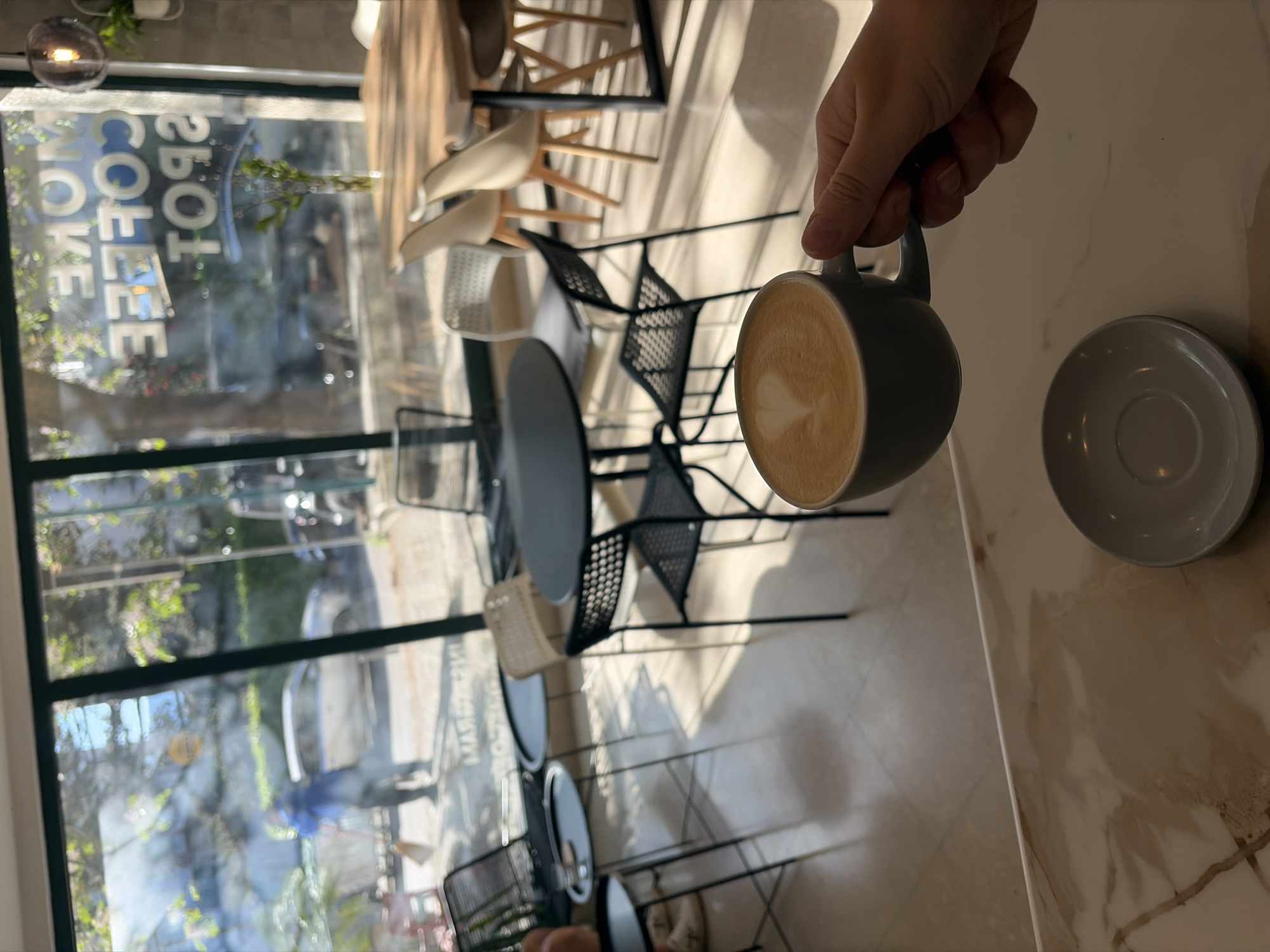 The photograph depicts a café interior with a partial view of an individual holding a cup of coffee above a saucer. The café features a window with the phrase "MORE SPOT COFFEE" printed in reverse. The tables and chairs are visible through the window and within the interior space. A hand holds the cup of coffee, indicating an action of serving or taking a drink. The environment appears to be a modern café setting. The location of Budva, Montenegro, cannot be confirmed or denied based on the image.