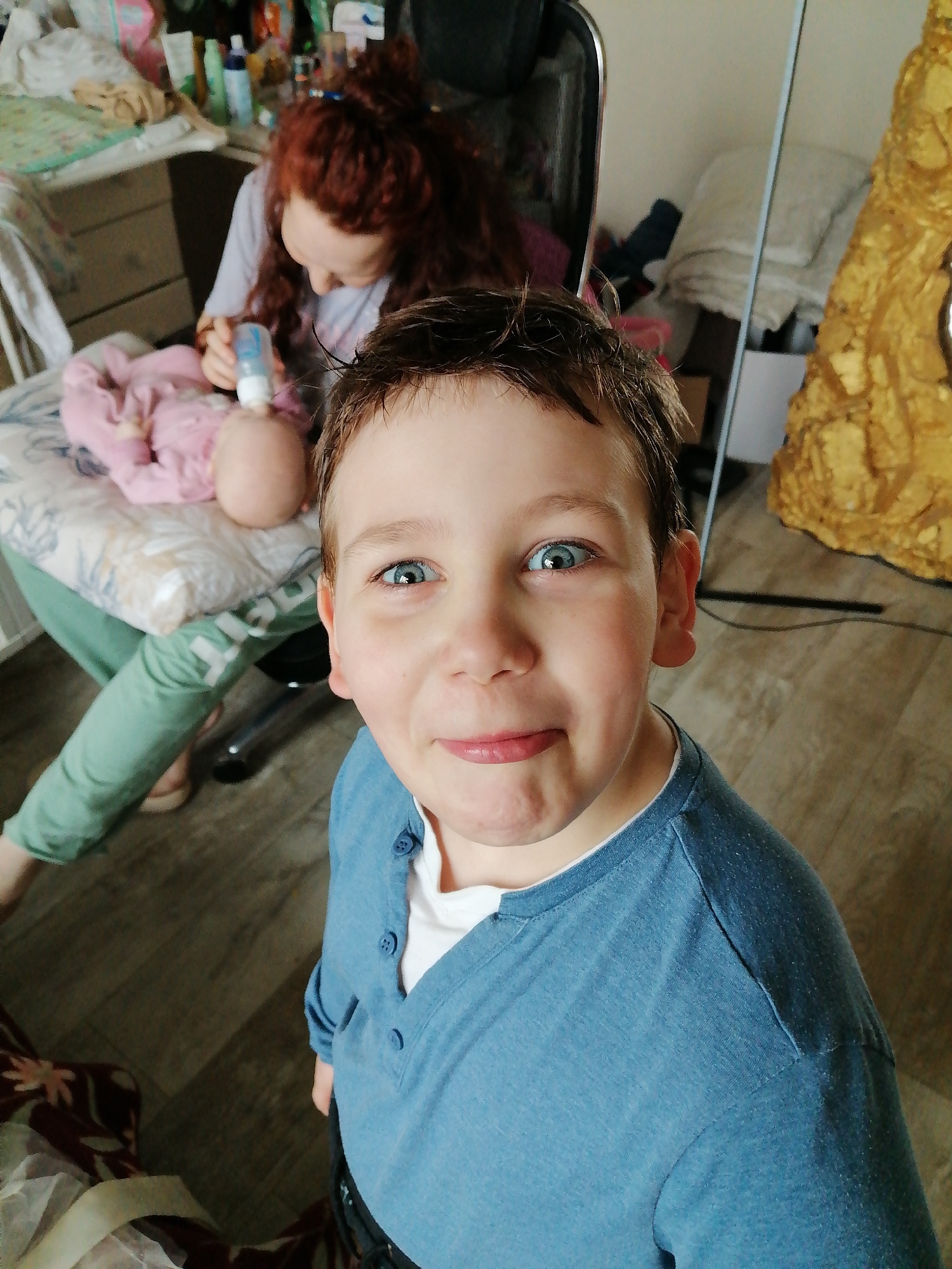 A close-up, slightly high-angle shot captures a young boy with fair skin, short brown hair, and striking blue eyes. He wears a blue collared shirt over a white undershirt, and his expression is a slight, subtle smile. The background features a woman with long, red, curly hair seated and holding a baby in pink clothing. She appears to be feeding the baby from a bottle. A table with various items is partially visible to the left. The floor is light brown and appears to be made of laminate or wood. A decorative gold object is visible to the right. The setting is indoors.