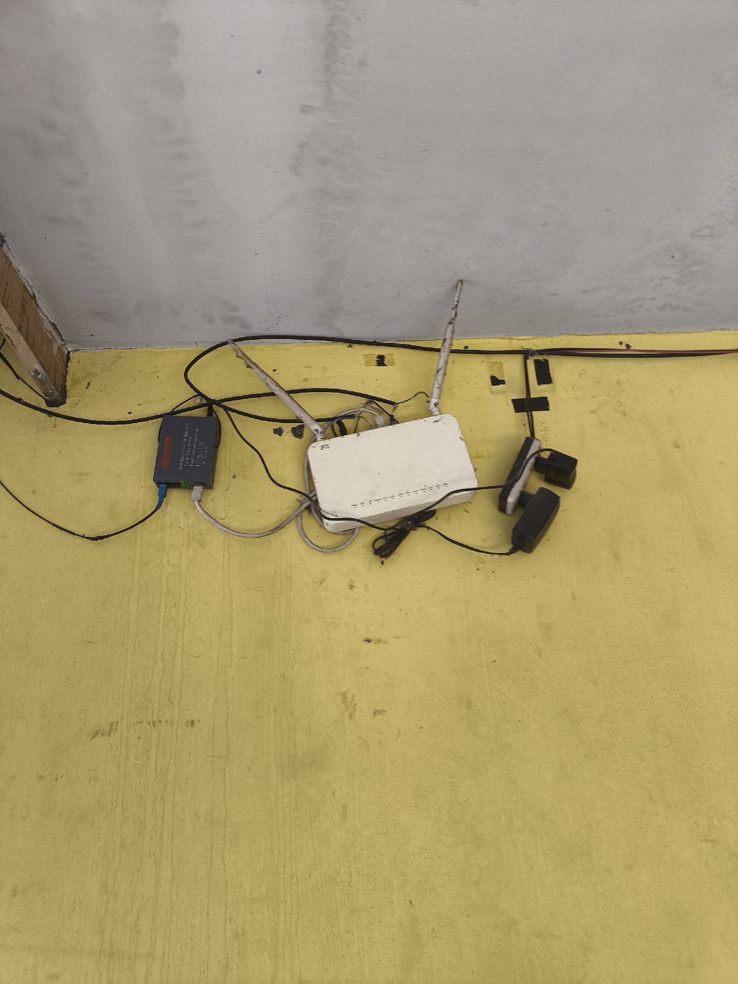 The image shows a white Wi-Fi router and a grey network device with various cables connected to them, mounted on a yellow wall. Two black power adapters are also visible, attached to the wall with black tape. The wall appears to be indoors and has a textured, possibly dirty or stained surface. A section of a concrete ceiling is visible at the top of the image. The overall scene suggests a functional but somewhat disorganized setup for internet connectivity, likely in a residential or small business setting. The image's origin is not specified, but it depicts common electronic equipment.