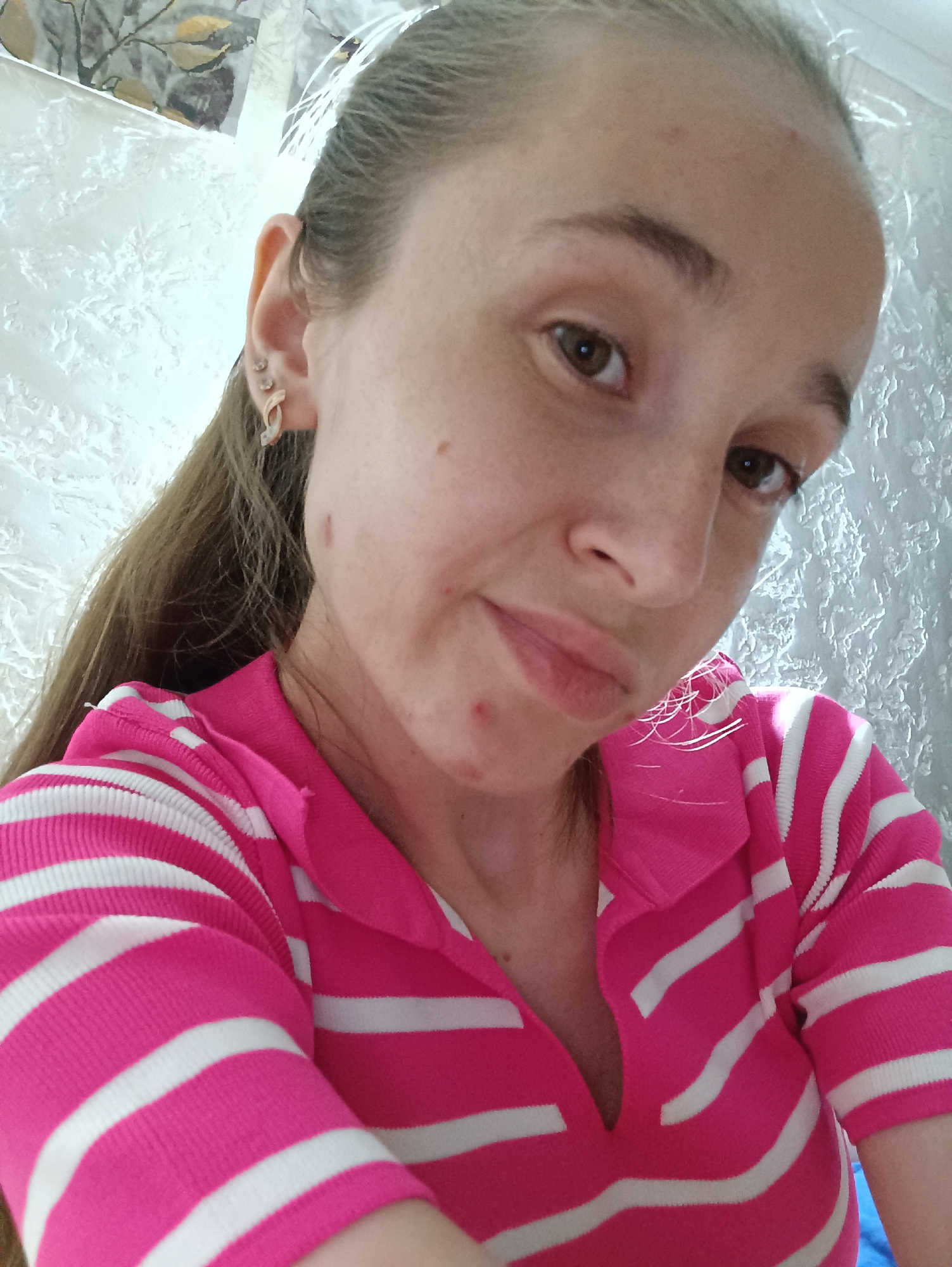The image is a close-up, selfie-style photograph of a young woman taken indoors. She has long, brown hair pulled back into a ponytail. She is wearing a pink and white striped polo shirt. Her skin appears fair with some visible blemishes. She has earrings in her left ear, including a hoop earring and stud earrings. The background appears to be a wall with a textured surface and some decorative elements, possibly a picture or tapestry. The lighting suggests an indoor setting, possibly a home. No specific location within Vypasne, Ukraine can be determined from the image itself.