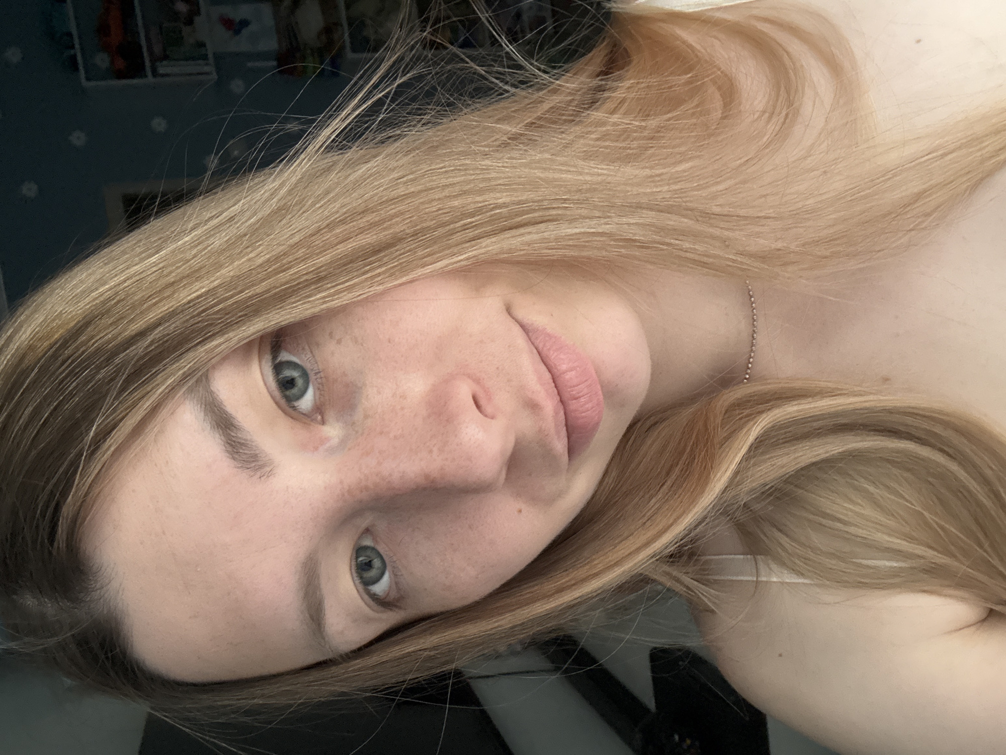 The image is a close-up selfie of a fair-skinned, young woman with blonde hair and blue eyes. She has freckles on her face. A thin silver necklace is visible. The background is out of focus but appears to show a wall with a floral pattern and some posters or pictures. There is no evidence of location within the city of Moscow, Russia.