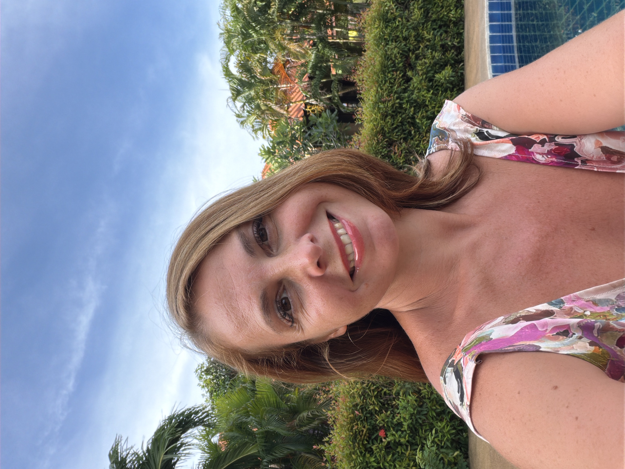 A woman with shoulder-length, light brown hair smiles for a selfie taken outdoors. She is wearing a floral-patterned top with shades of pink, purple, white, and green. Her skin is lightly tanned, and she has freckles visible on her shoulders and arms. Behind her, lush green foliage, including palm trees and hedges, forms a backdrop. In the lower right corner of the image, a portion of a blue-tiled swimming pool is visible. The sky above is a clear blue with a few wispy white clouds. The scene appears to be a resort or a residential area in a tropical location, potentially Baan Mae Nam, Thailand, given the vegetation and climate suggested by the image.
