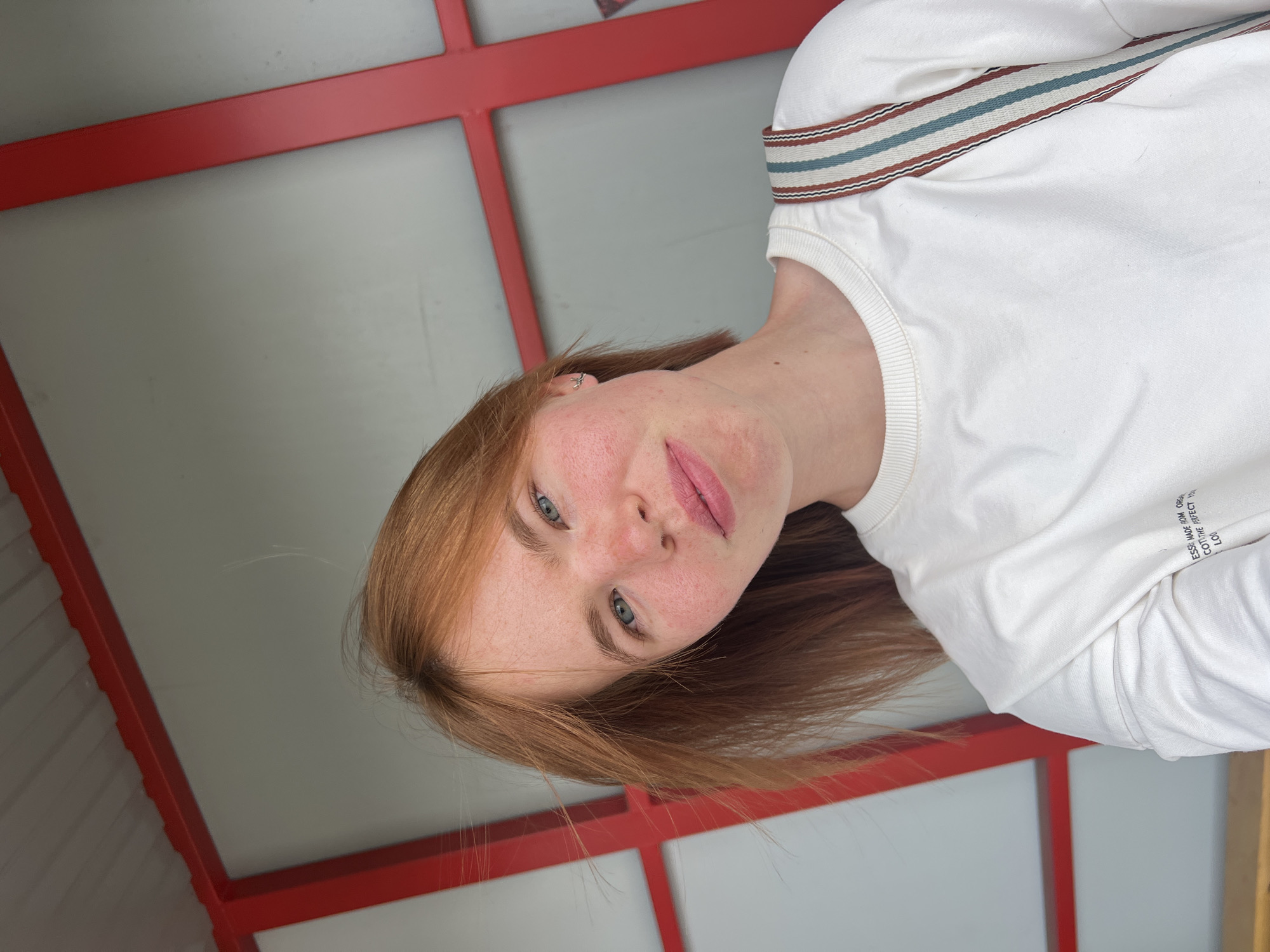 A young woman with auburn hair and fair skin, sporting a light dusting of freckles, is pictured from the chest up, looking towards the camera. She has blue eyes and a gentle smile. She is wearing a white crew-neck sweatshirt. A bag strap with red, blue, and white stripes is visible over her right shoulder. The background consists of a light gray wall framed by a red structure, possibly part of an outdoor pavilion or architectural feature. The image appears to have been taken outdoors, likely in a public space within the city of Chaikovsky, Russia. The lighting suggests it is daytime.