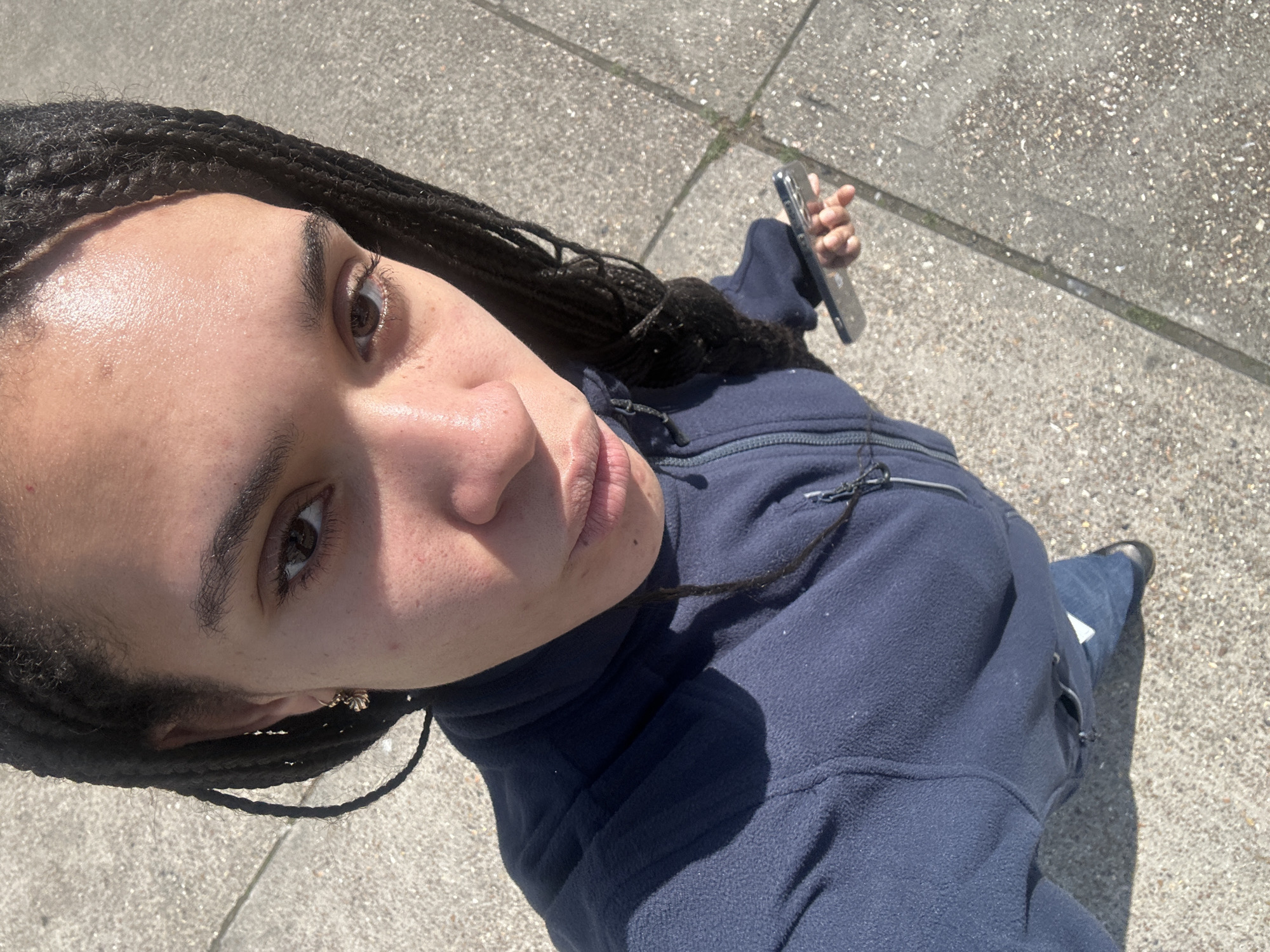 This is a top-down, slightly rotated outdoor selfie featuring a young adult with dark, braided hair. The individual looks directly upwards at the camera with a neutral to slightly contemplative expression. Their right eye is prominent, revealing a dark iris, with portions of their nose and lips also visible. Their skin appears reflective, possibly due to the bright sunlight or sweat, and shows a few minor blemishes or freckles. A small, possibly gold-colored earring is visible in their left ear.
The person is wearing a dark blue fleece jacket with a high collar and visible zipper and drawstring details. Their left arm, clad in a blue jean, is partially visible, leading down to a dark shoe. In their right hand, they casually hold a silver smartphone.
The setting is an outdoor paved area, likely a sidewalk or urban path, composed of light grey concrete with visible aggregate. Strong, distinct shadows are cast from the person's body onto their jacket and the ground, indicating bright, direct sunlight, suggesting a clear day during midday or early afternoon. While the image itself lacks specific visual identifiers, the context provided states the location is London, United Kingdom. No text is visible within the image.