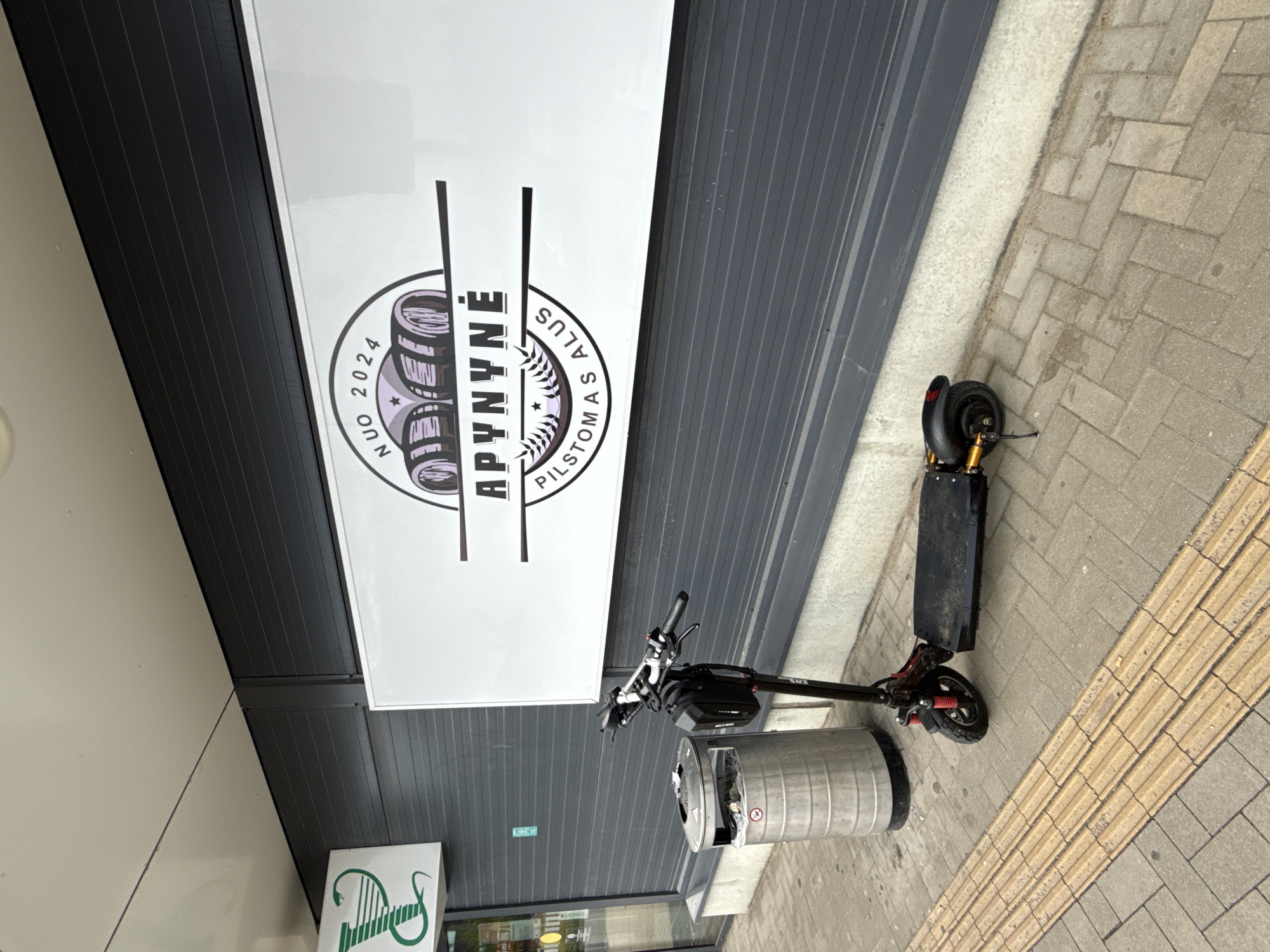 An electric scooter is parked on a sidewalk in front of a building with a large sign. The scooter is black with red accents and has a black bag attached to the handlebars. To the left of the scooter is a silver trash can. The building has dark gray siding and a white sign with black lettering and graphics. The sign appears to be an advertisement. The sidewalk is paved with gray and yellow rectangular tiles. A portion of a green sign with a white symbol is visible on the left side of the building. The weather appears to be overcast.